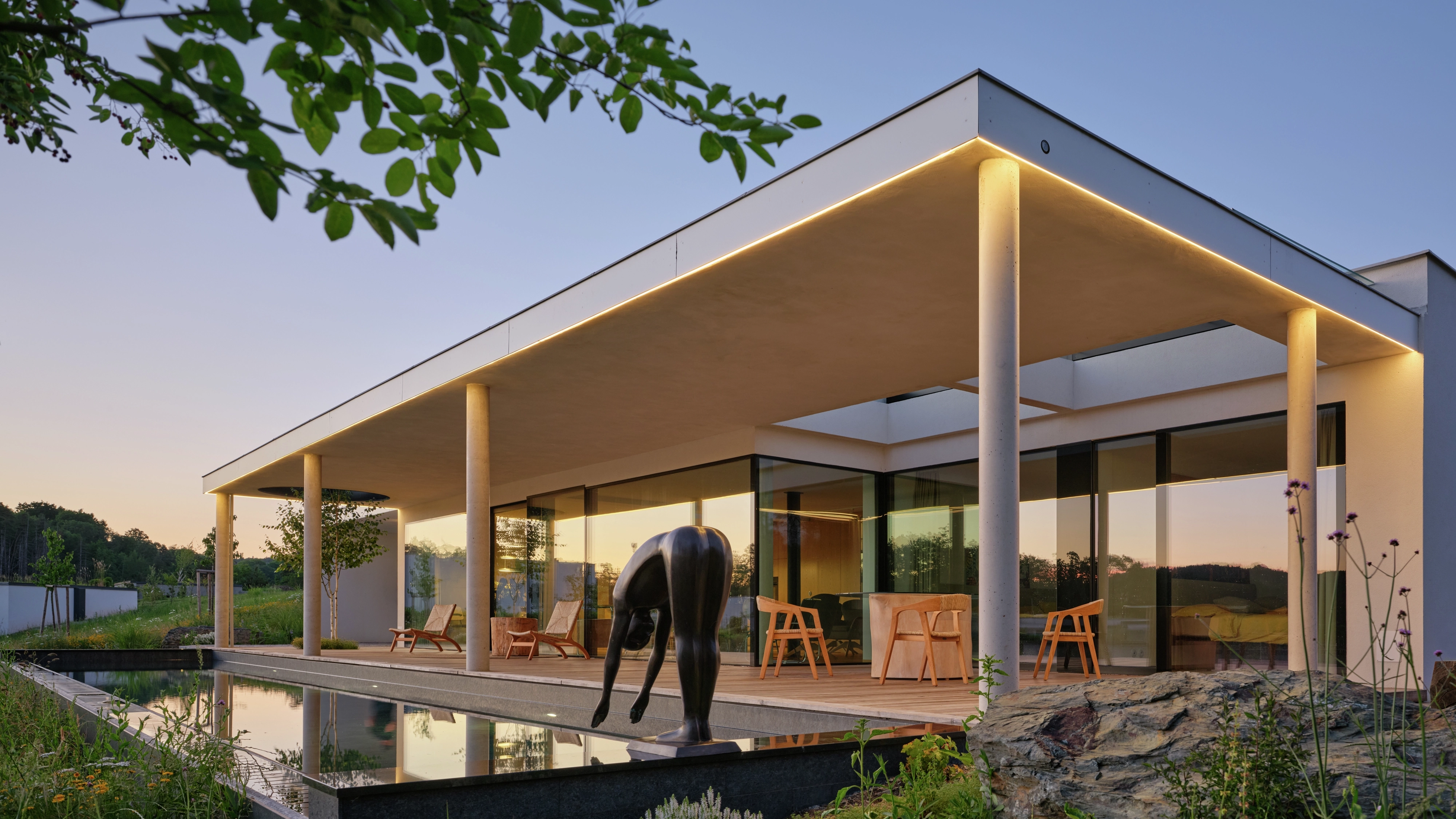Modern flat-roof house with glass walls, terrace, and reflecting pool at dusk.