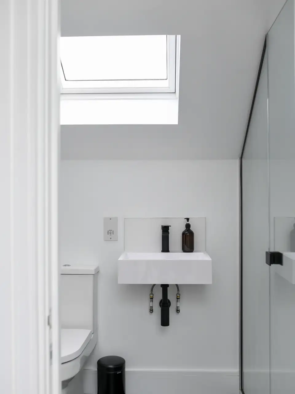 Bagno minimal di colore bianco con doccia vetrata illuminato da una finestra per tetti.