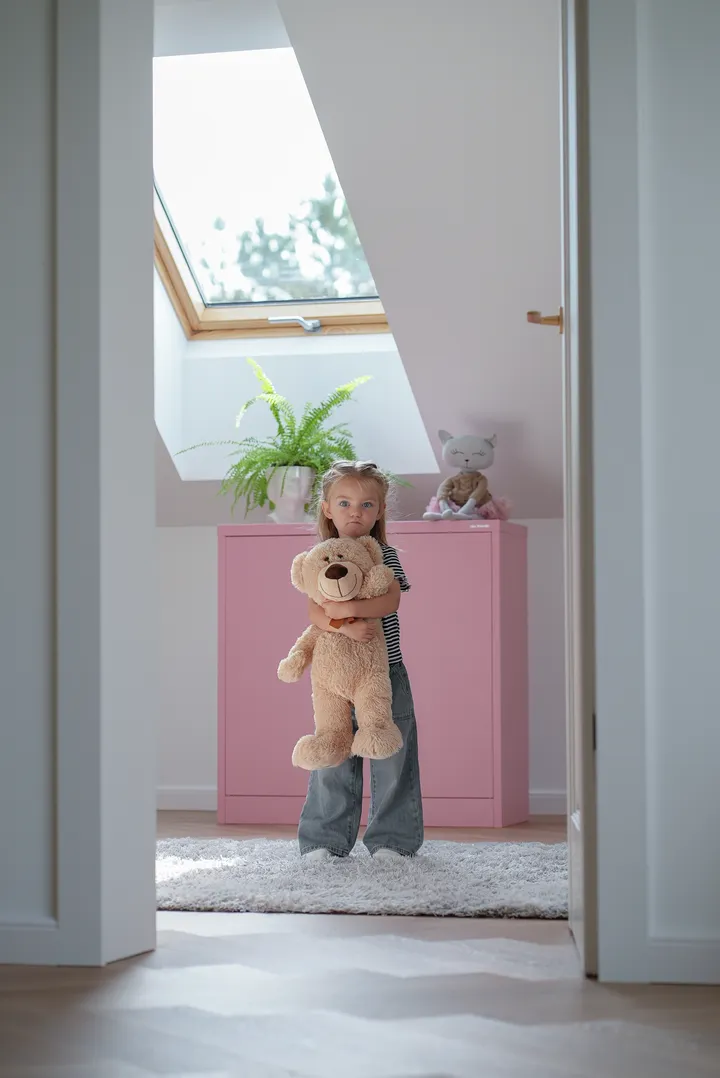 Dziewczynka trzymająca pluszowego misia pod oknem dachowym w jasnym pokoju na poddaszu | VELUX