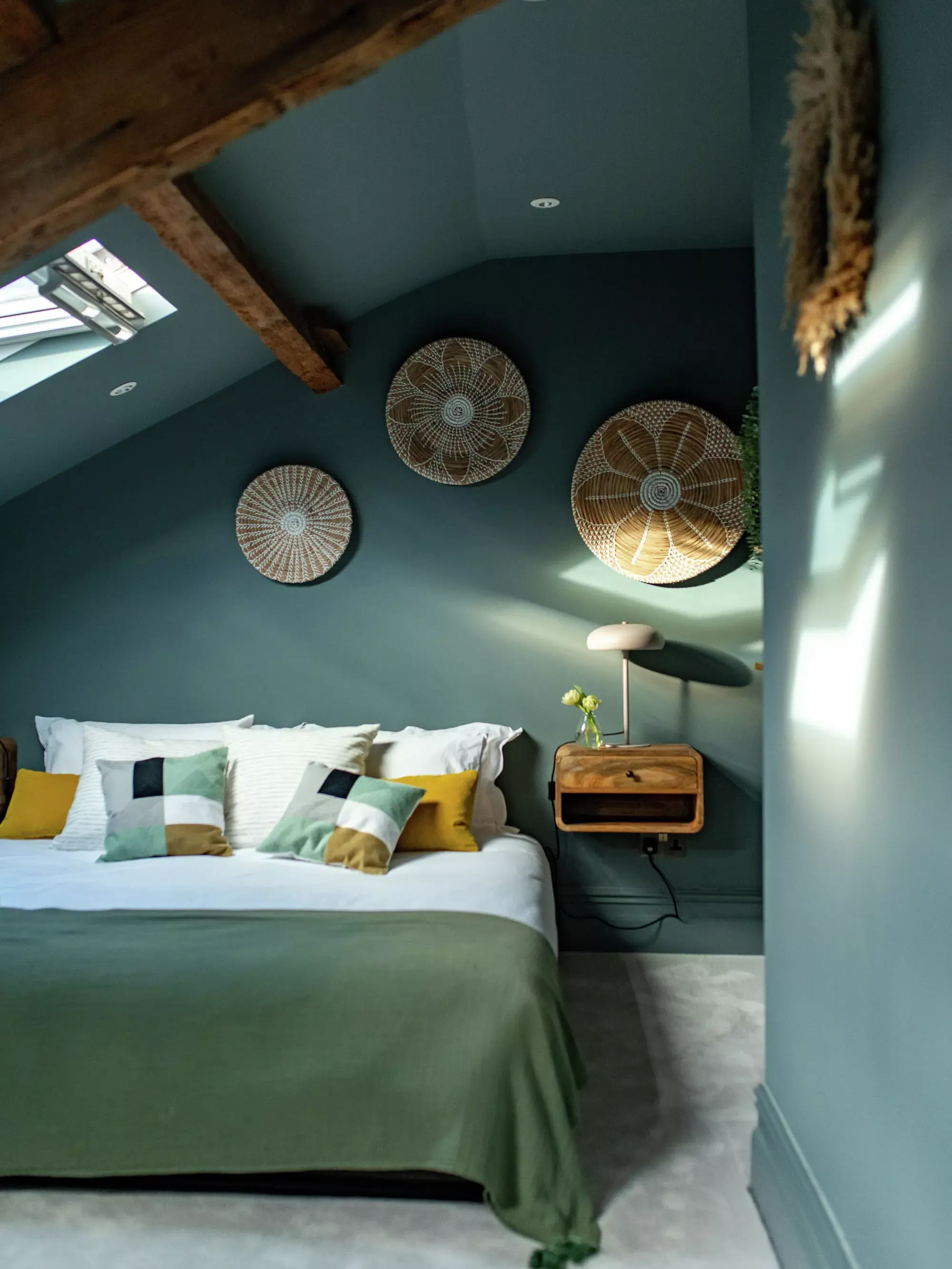 Chambre sous combles avec lit vert, coussins décoratifs, et paniers muraux en rotin