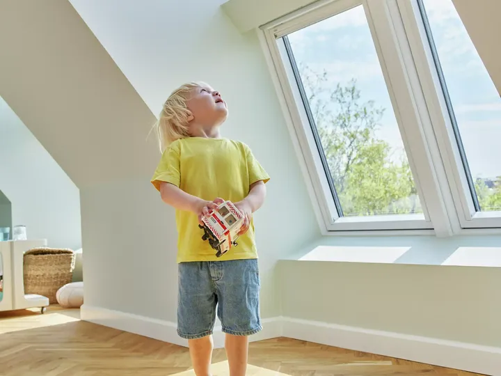 Kind in einem sonnendurchfluteten Dachbodenraum mit einem VELUX Dachflächenfenster und hölzernem Boden, das ein Spielzeug hält.