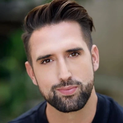Close-up of a young man with dark hair and beard looking at the camera outdoors