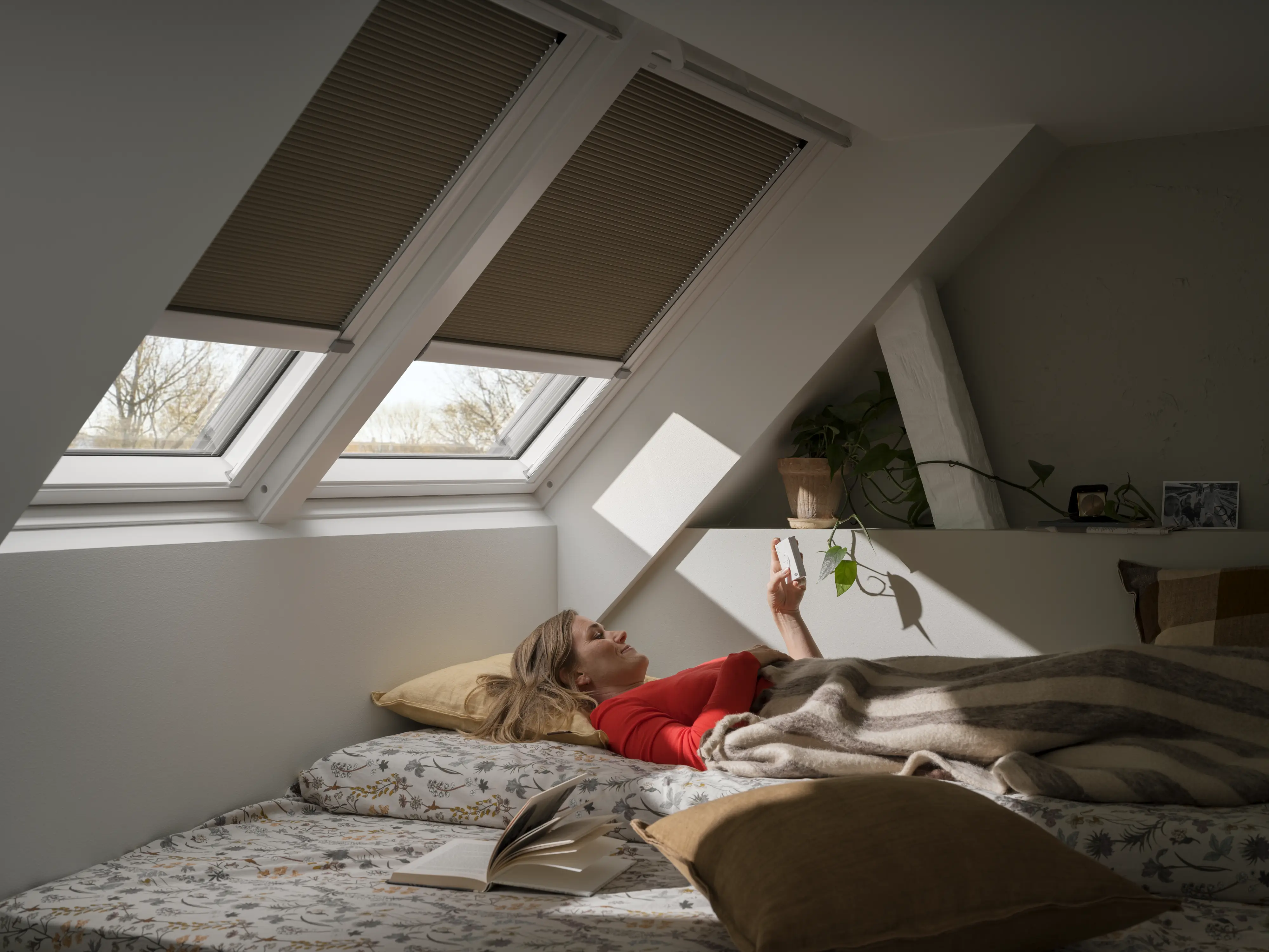Nő pihen tetőtéri hálószobában, ahol a VELUX fényzáró pliszé árnyékolók szabályozzák a bejutó fényt, és segítenek egyensúlyt teremteni a bevilágítás és a sötétítés között.