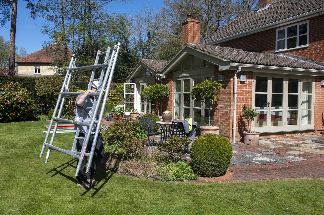 Man carrying a ladder in a garden with a brick house in the background.