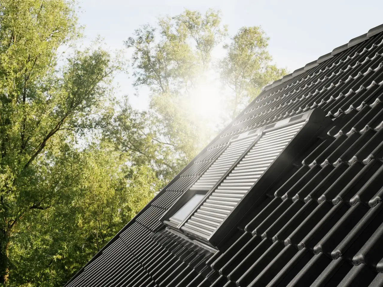 Roof with black  tiles and closed VELUX roof windows with solar shutters, surrounded by trees.