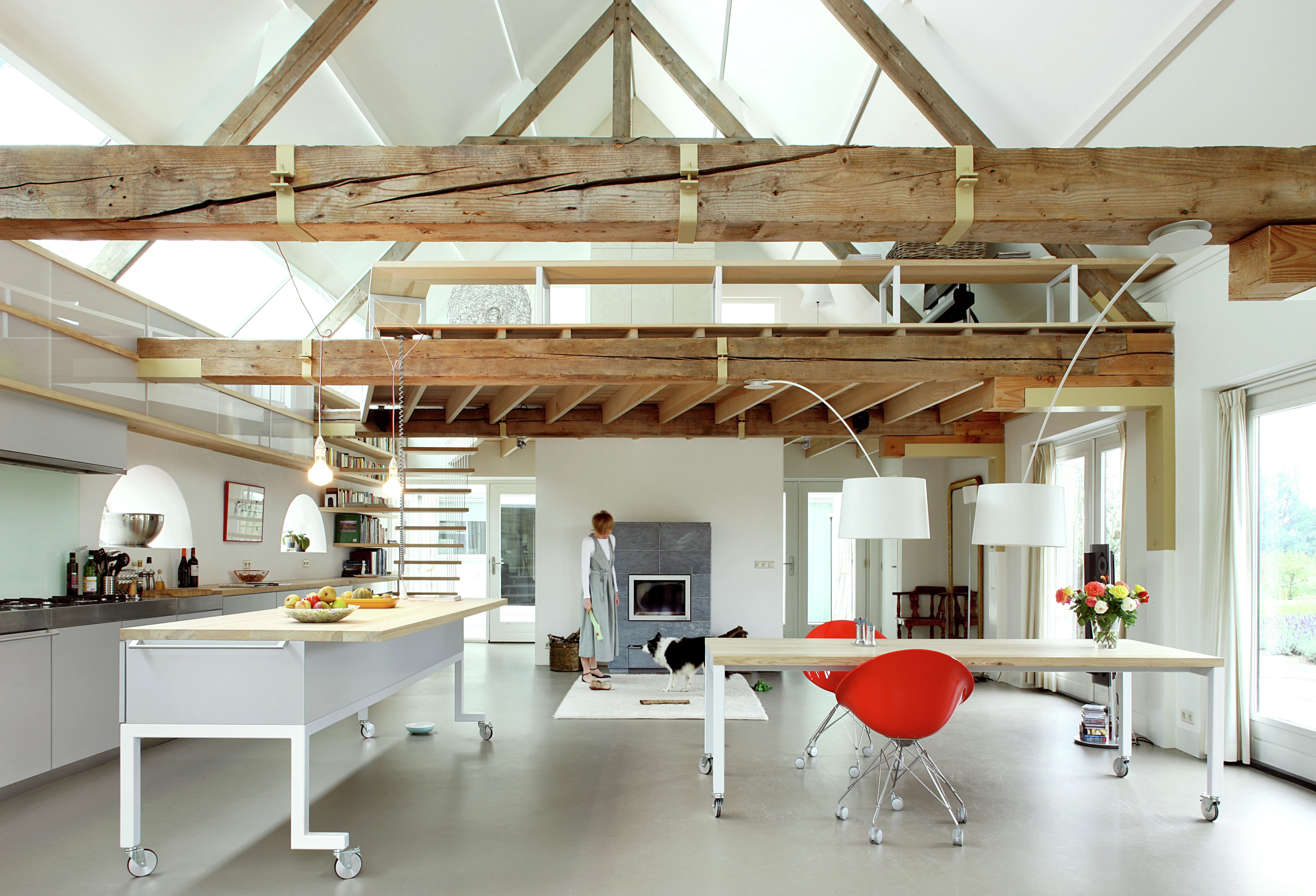 Interno di un’ampia e moderna mansarda con travi di legno a vista. La stanza presenta un’area cucina con mobili bianchi e un’isola centrale, un tavolo da pranzo con sedie e uno spazio living con una sedia a bolla rossa sospesa. Grandi finestre permettono alla luce naturale di riempire lo spazio, evidenziando il contrasto tra gli arredi contemporanei e il fascino rustico della struttura in legno.