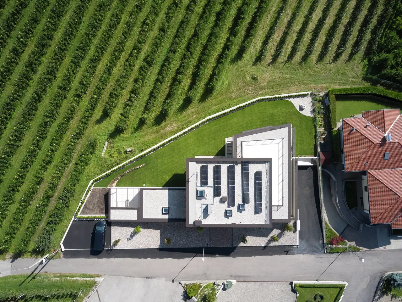 Vue aérienne d’une maison moderne avec un toit plat, entourée d’une pelouse bien entretenue et adjacente à des rangées de cultures ou de vignobles. La maison est située à côté d’une route avec un autre bâtiment au toit de tuiles rouges.