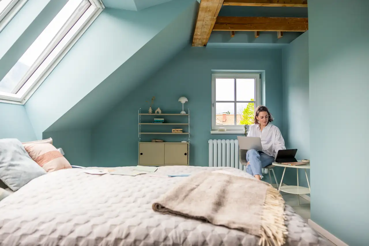 Aesthetic-Schlafzimmer in Blautönen mit Bett unter Dachfenstern und Frau am Laptop | VELUX Magazin
