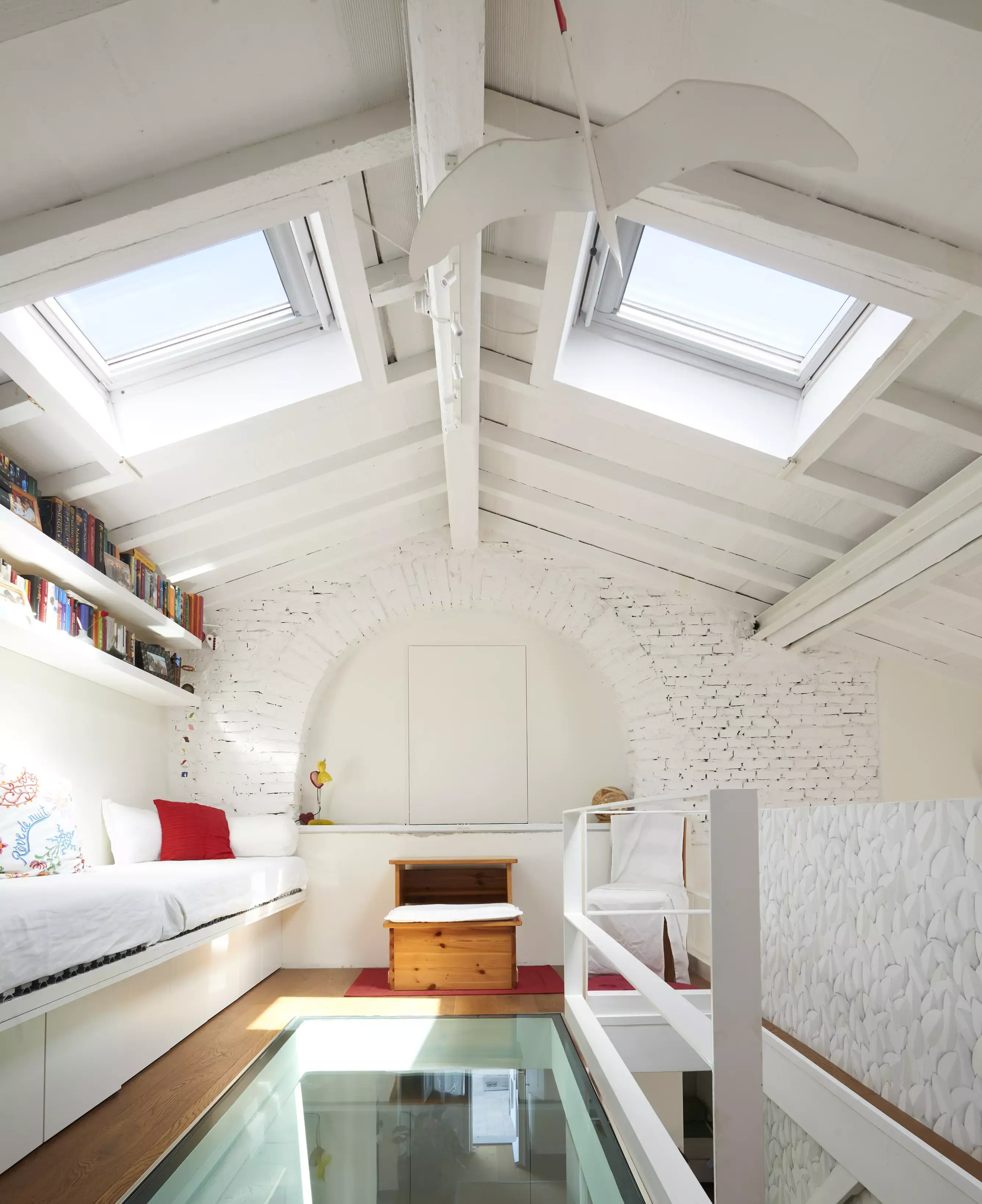 Camera da letto mansarda con finestre per tetti VELUX, muri di mattoni bianchi e dettaglio del pavimento in vetro.
