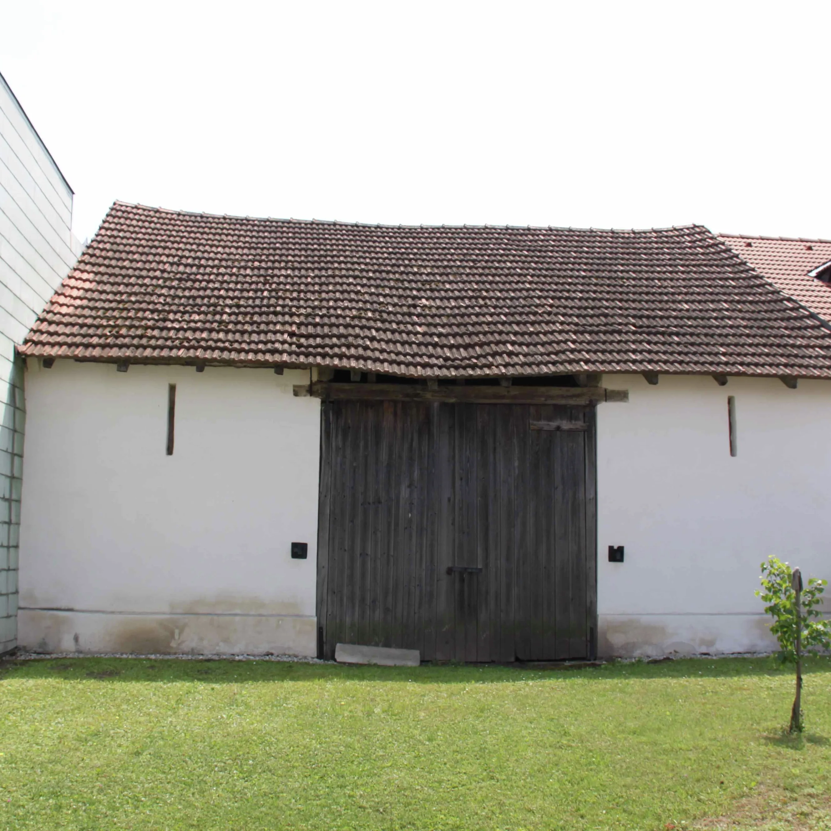 Rustic barn with tiled roof and wooden door, surrounded by modern buildings.