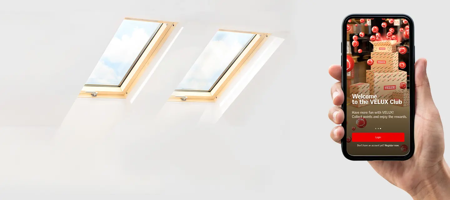 Two VELUX roof windows in a bright attic beside a hand holding the VELUX Club app on a smartphone.