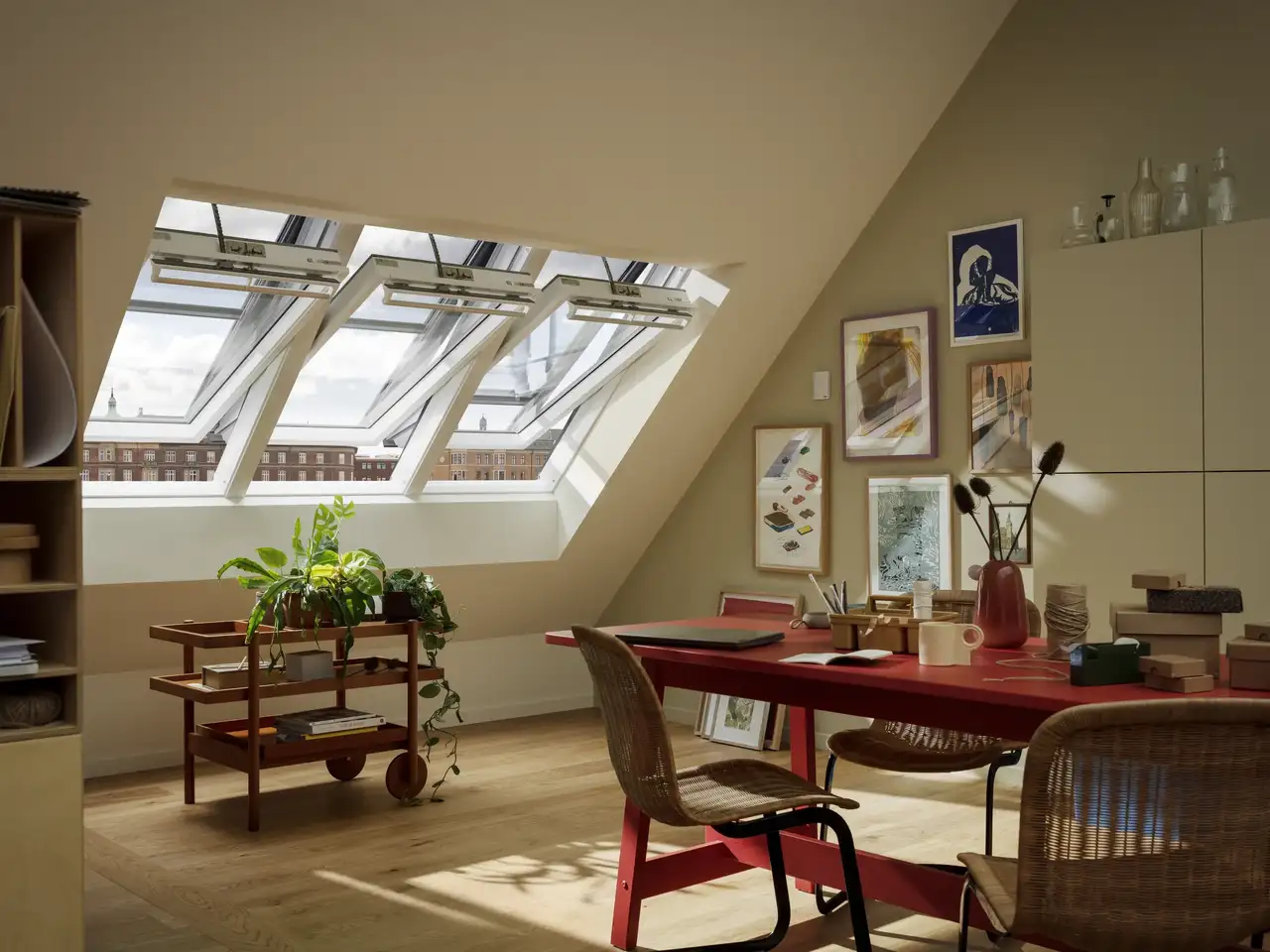 Delovni prostor v mansardi s poševnim stropom in odprtimi strešnimi okni VELUX, umetninami in rastlinami.