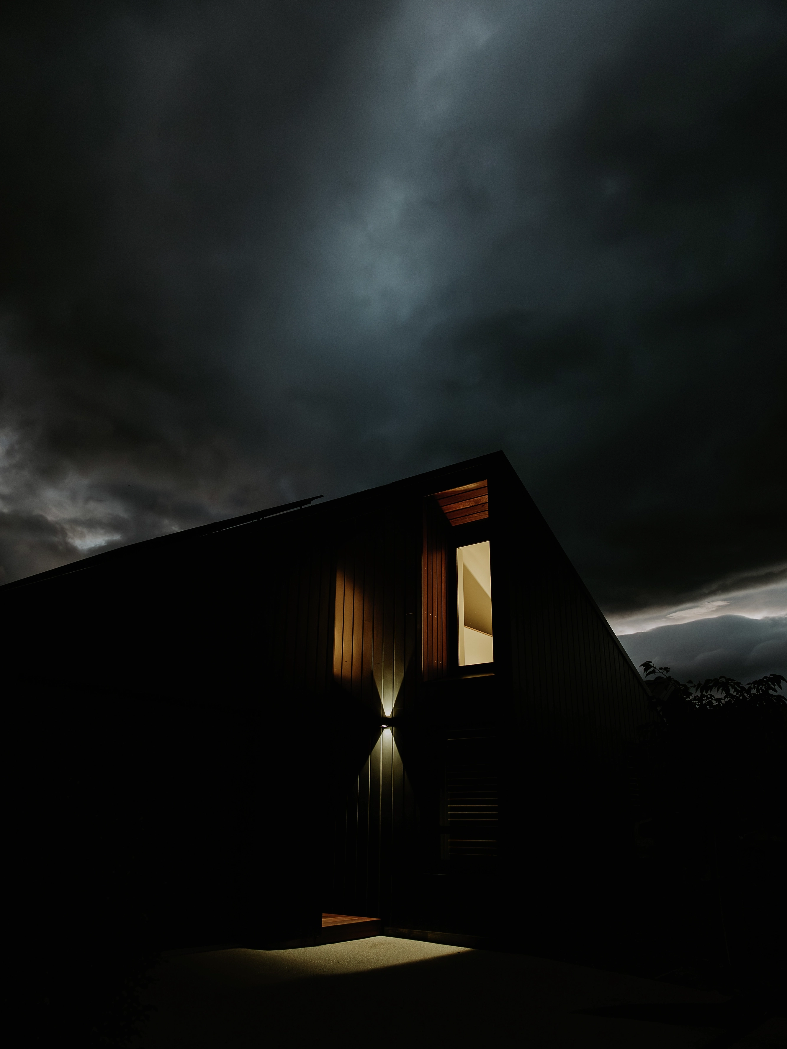 Modern dark-clad house at night with one lit upper window under stormy clouds.