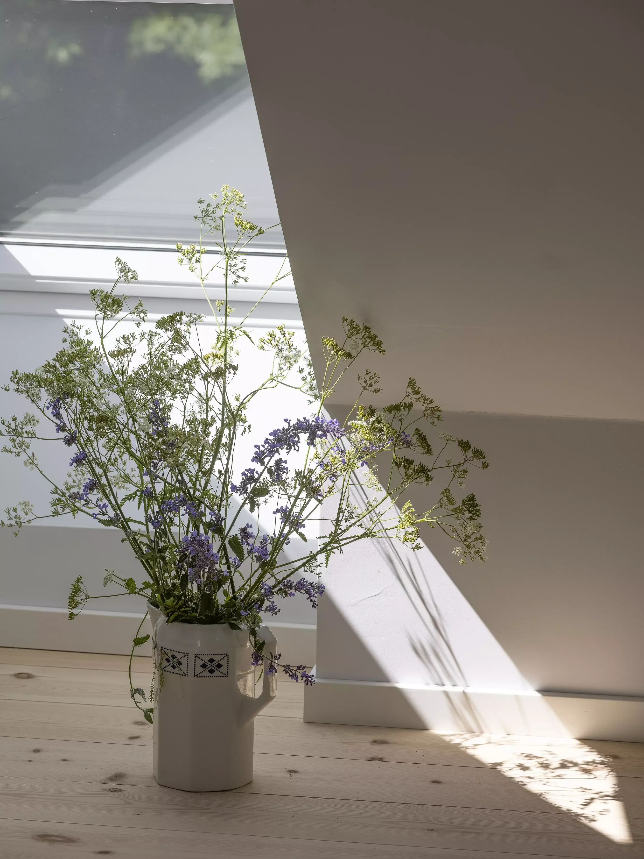 Lumière du soleil à travers une fenêtre VELUX dans le grenier, projetant des ombres sur le sol avec un pichet rempli de fleurs.