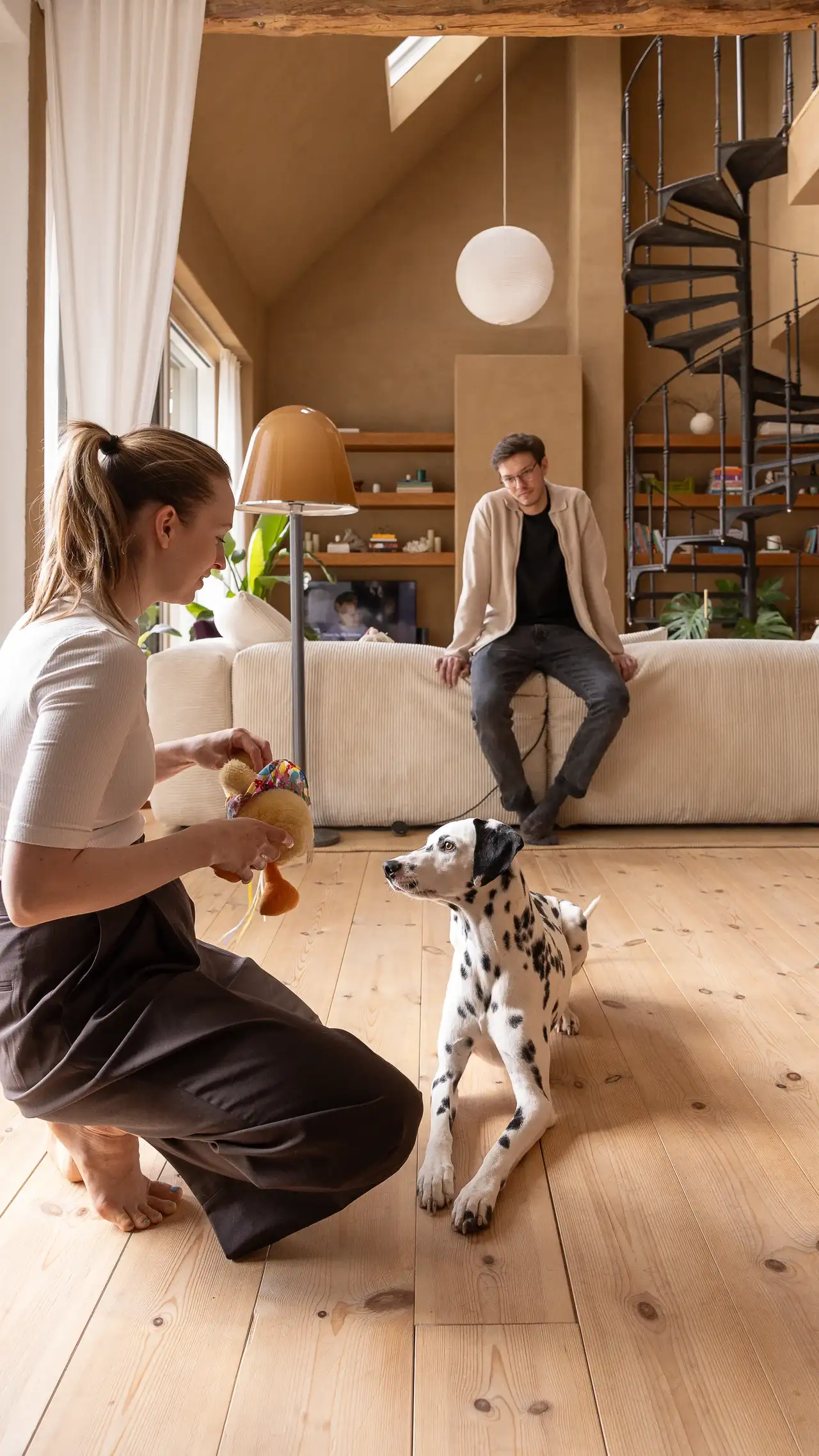 Eine Frau spielt mit ihrem Dalmatiner in einem sonnendurchfluteten Wohnzimmer, während ein Mann vom Sofa aus zusieht. VELUX Dachfenster bringen Tageslicht in den Raum. | VELUX Magazin