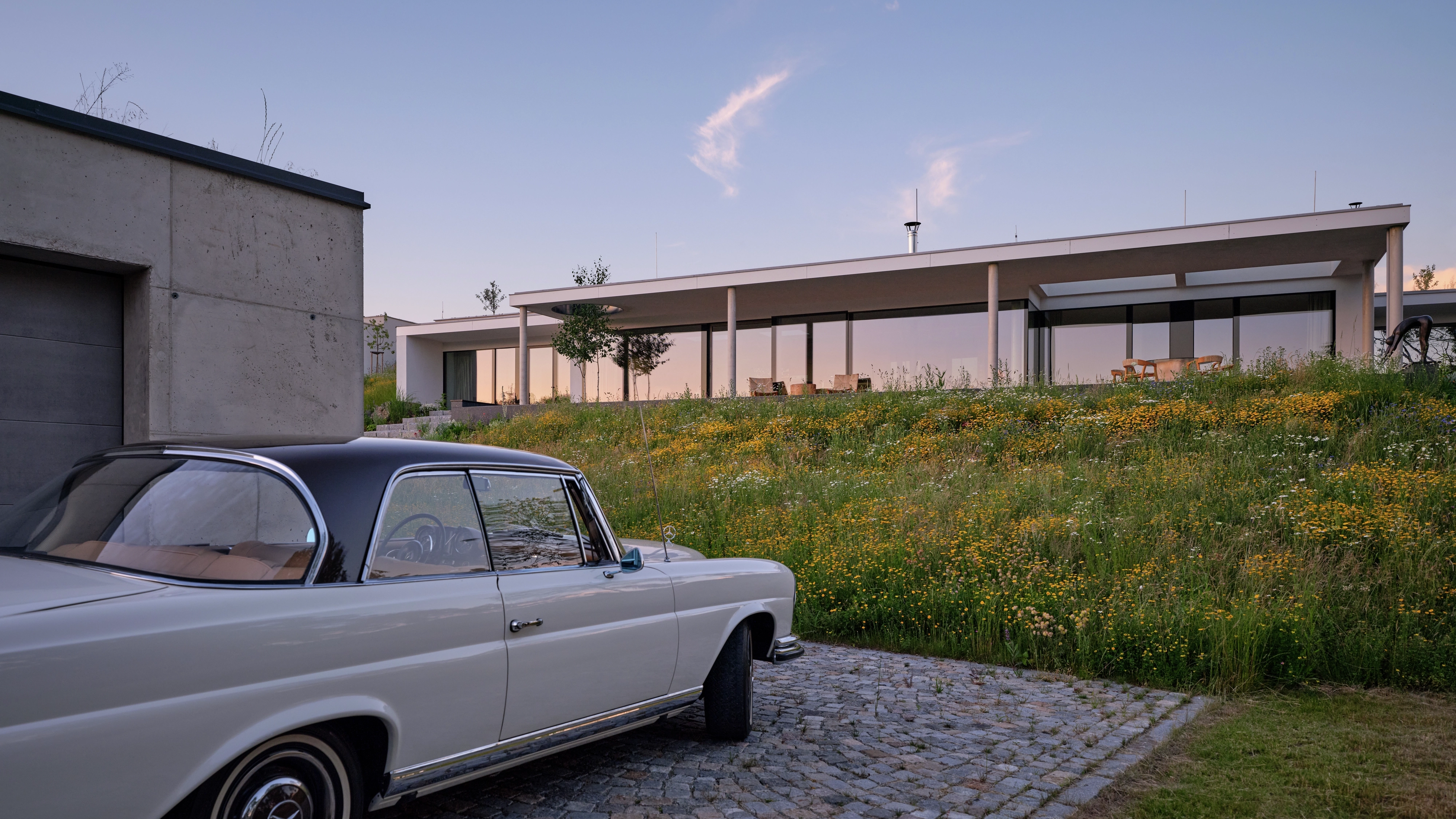 Modern flat-roof villa above meadow with vintage white car on cobblestone drive at sunset.