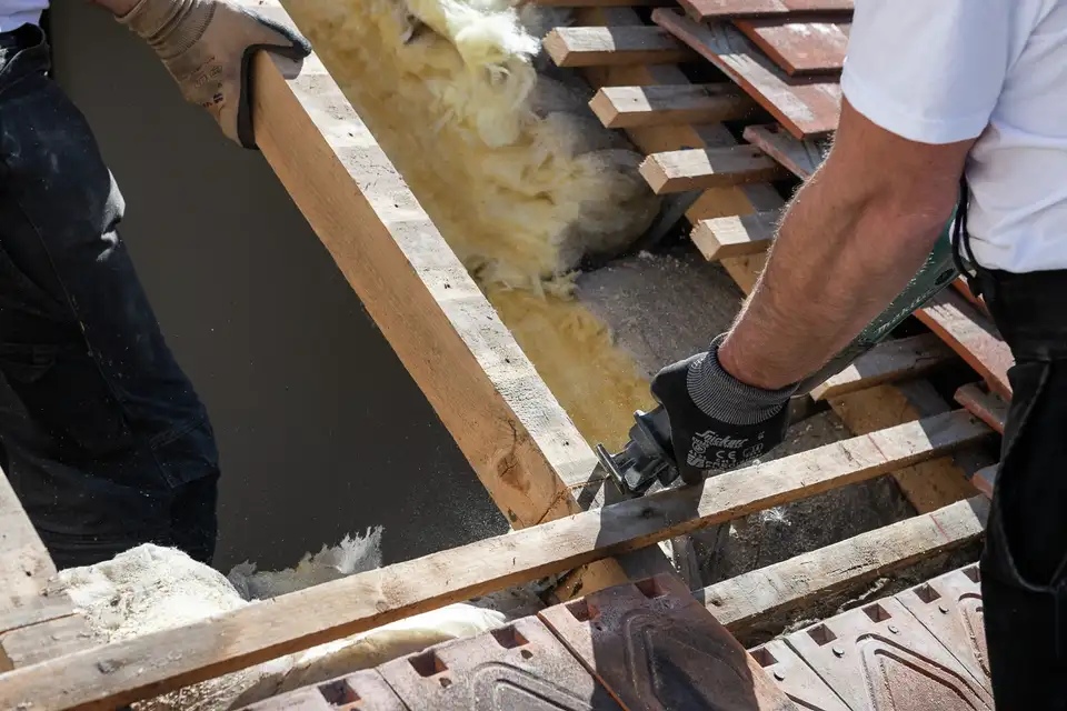 Trabalhadores a reparar vigas do telhado e isolamento, utilizando ferramentas e luvas. 
