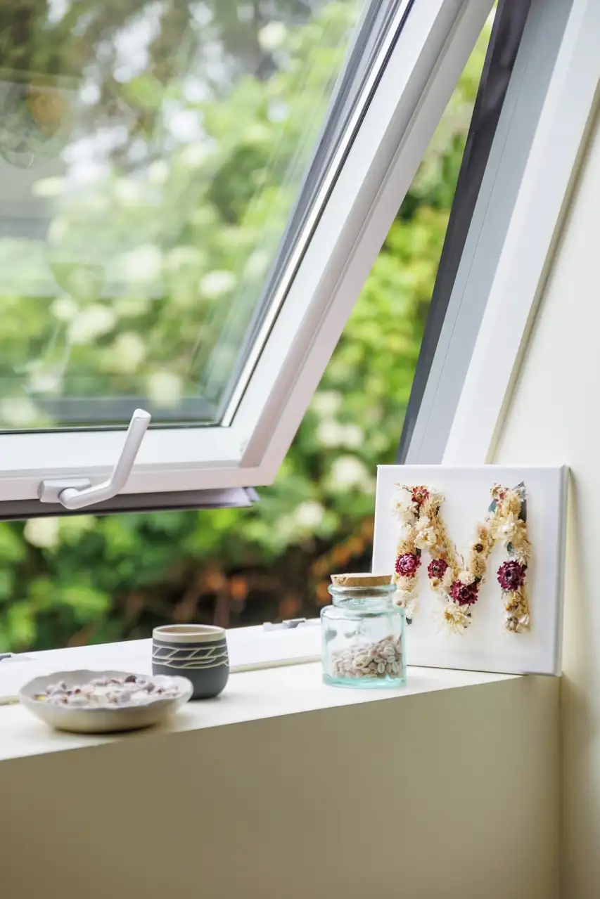 Une fenêtre de toit VELUX partiellement ouverte avec un rebord de fenêtre décoratif comprenant des bocaux, une petite toile et une vue sur la verdure. 