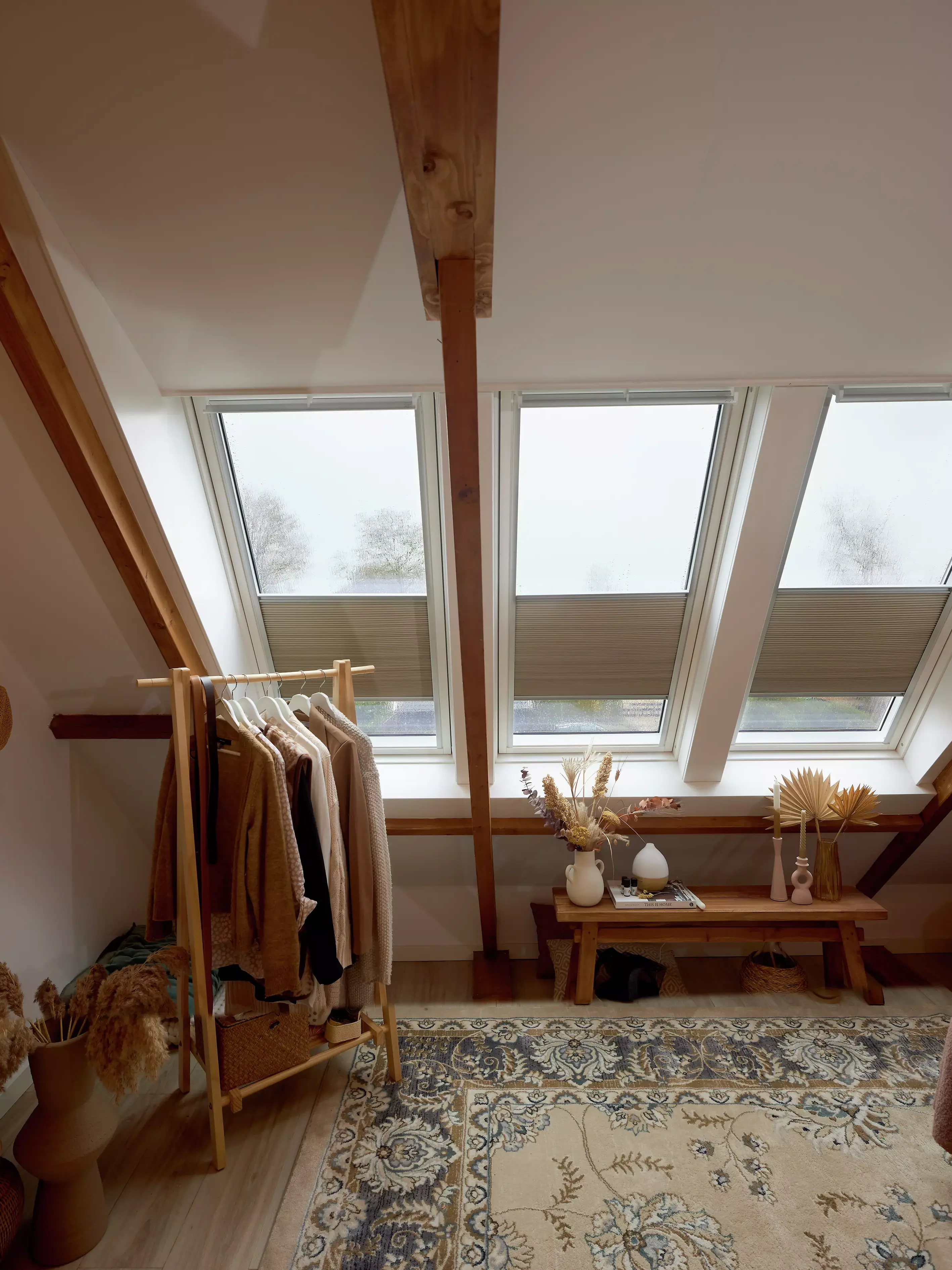 Espace de dressing dans le comble avec lumière naturelle provenant des fenêtres de toit VELUX, poutres en bois, et un tapis à motifs.