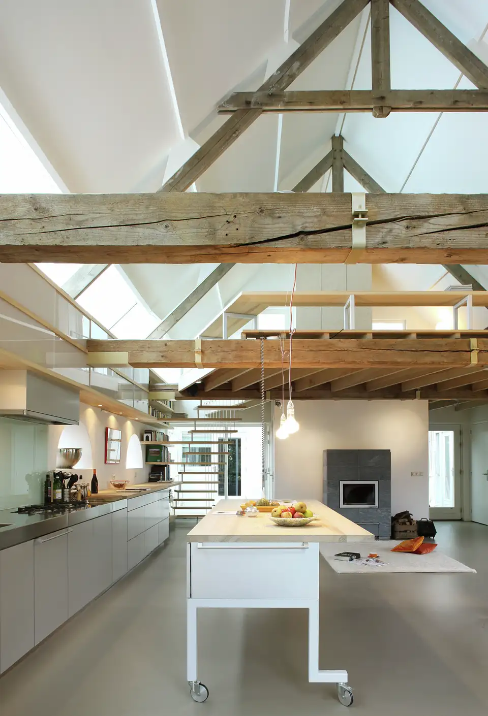 L’immagine mostra uno spazio interno con un soffitto alto caratterizzato da travi di legno a vista. Sotto le travi, c’è un livello mezzanino con una ringhiera metallica, e sotto di esso c’è un’area cucina con elettrodomestici moderni e un’isola centrale. Lo spazio ha grandi finestre che permettono un’abbondante luce naturale. Questa immagine è interessante in quanto mette in mostra un design architettonico che combina elementi rustici con uno stile contemporaneo, creando uno spazio abitativo aperto e arioso.