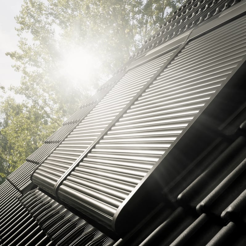 Roof with integrated solar panels reflecting sunlight, surrounded by trees.