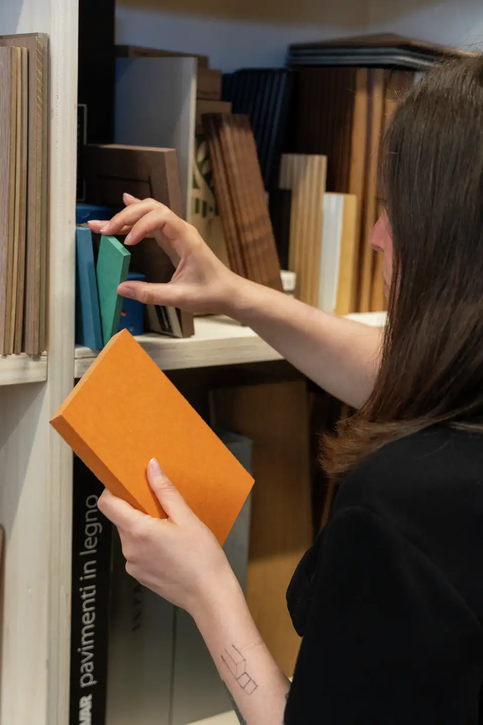 L’immagine mostra una persona che seleziona un libro da uno scaffale. La persona tiene un libro arancione in una mano e sembra stia cercando di prendere un altro libro con l’altra mano. Questa immagine potrebbe essere rilevante o interessante per discussioni sulla lettura, biblioteche, educazione o organizzazione personale.
