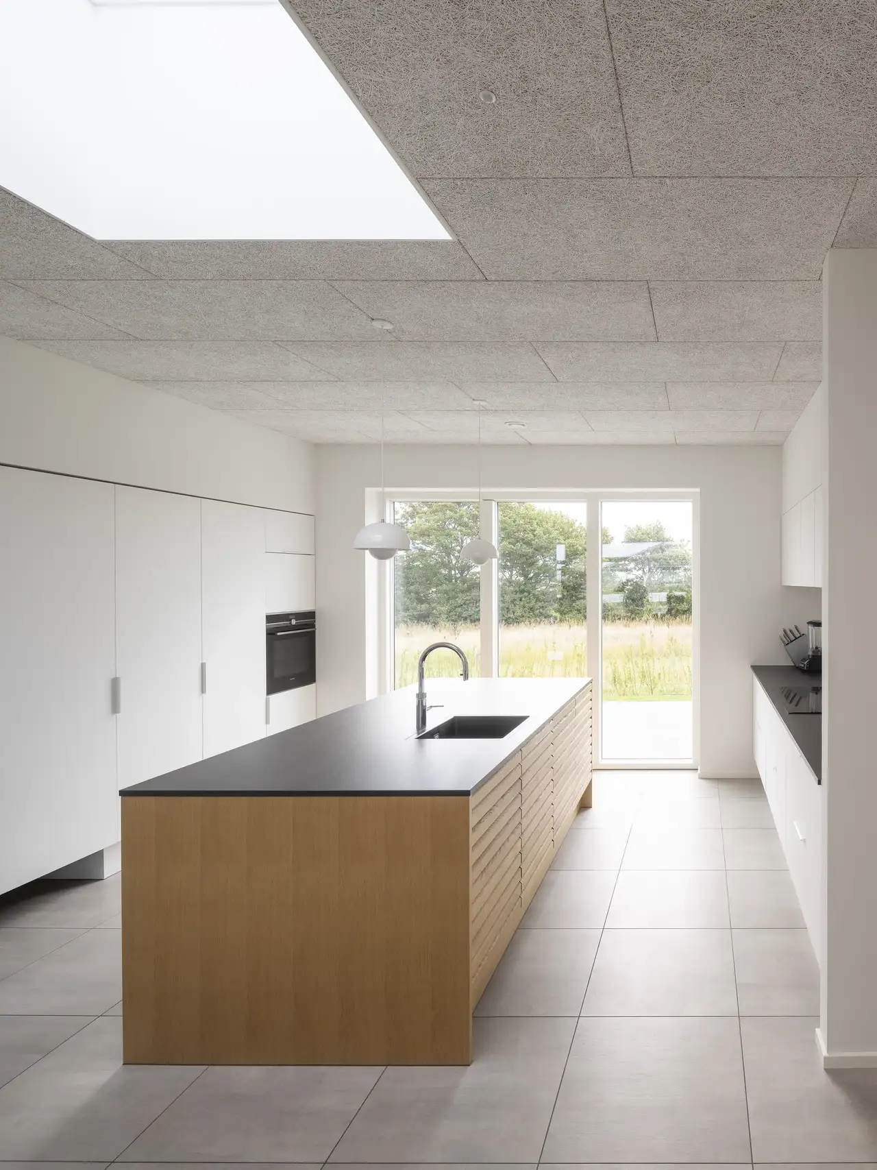 Cuisine blanche avec îlot en bois et plan de travail noir, éclairée par de grandes fenêtres verticales et une fenêtre pour toit plat VELUX.