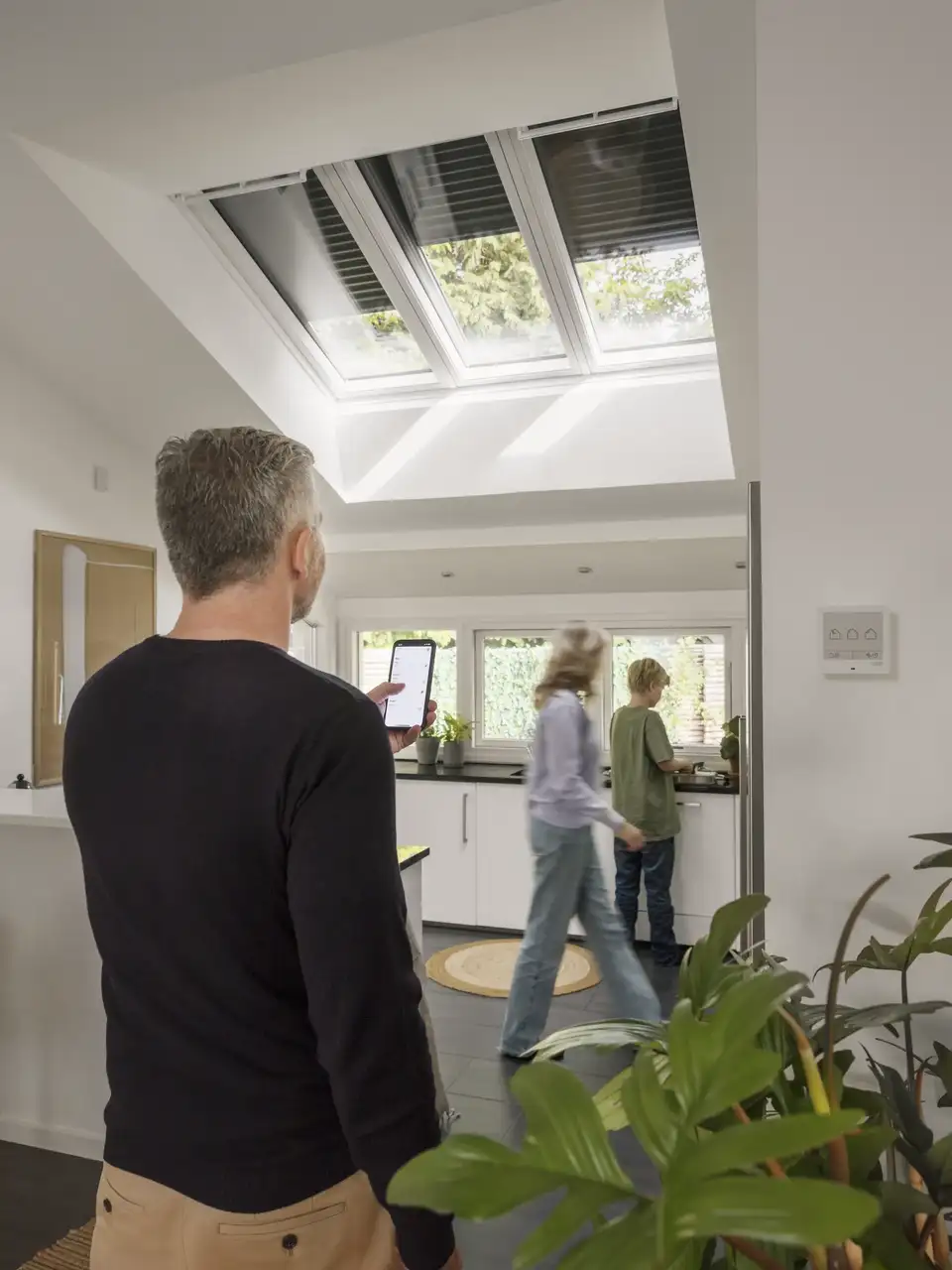 Un homme contrôle les fenêtres de toit et les volets roulants VELUX de sa cuisine à l’aide de l’application VELUX sur smartphone