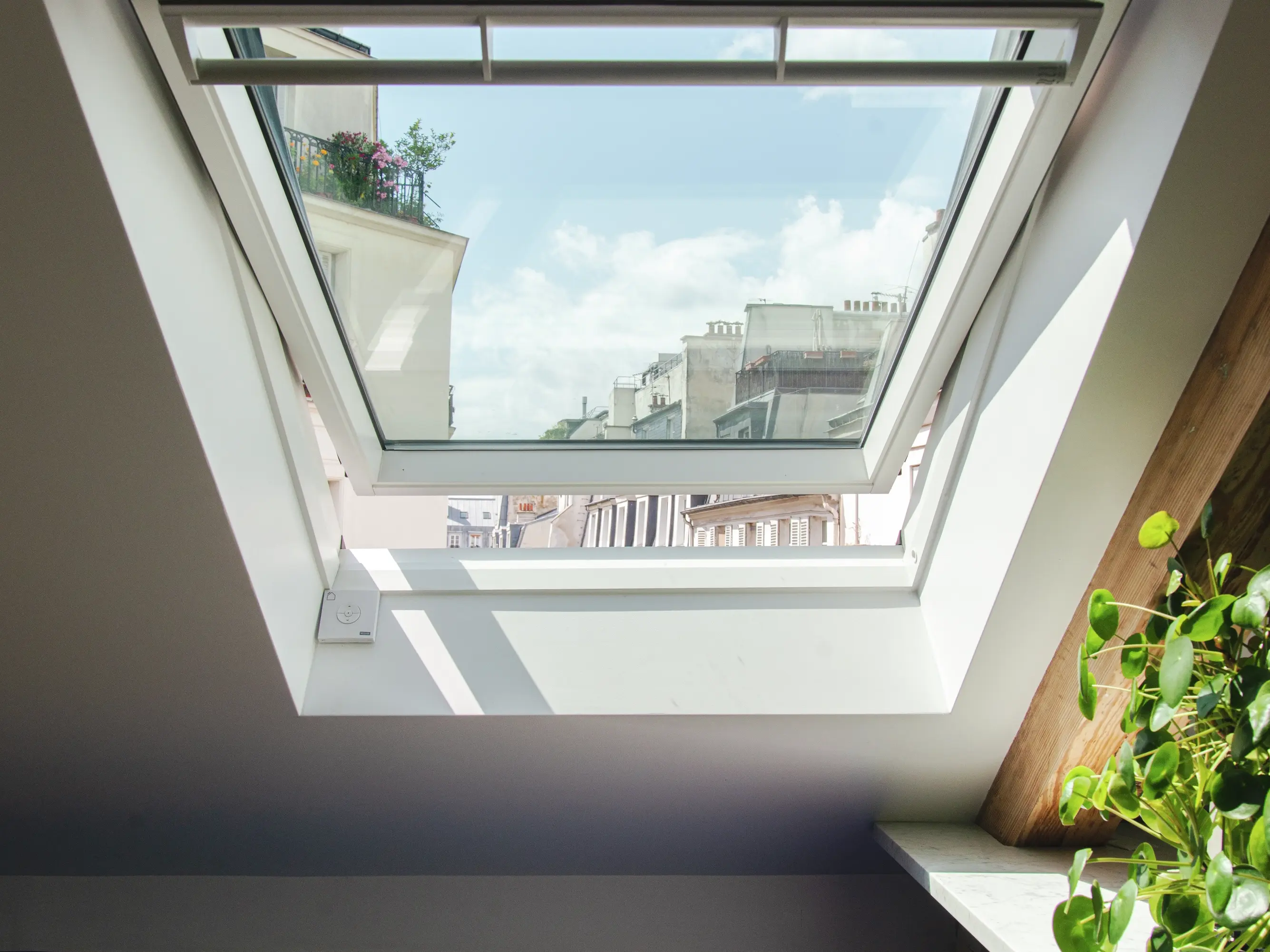 Fenêtre de toit VELUX dans la chambre du comble avec vue sur le ciel bleu et le paysage urbain.
