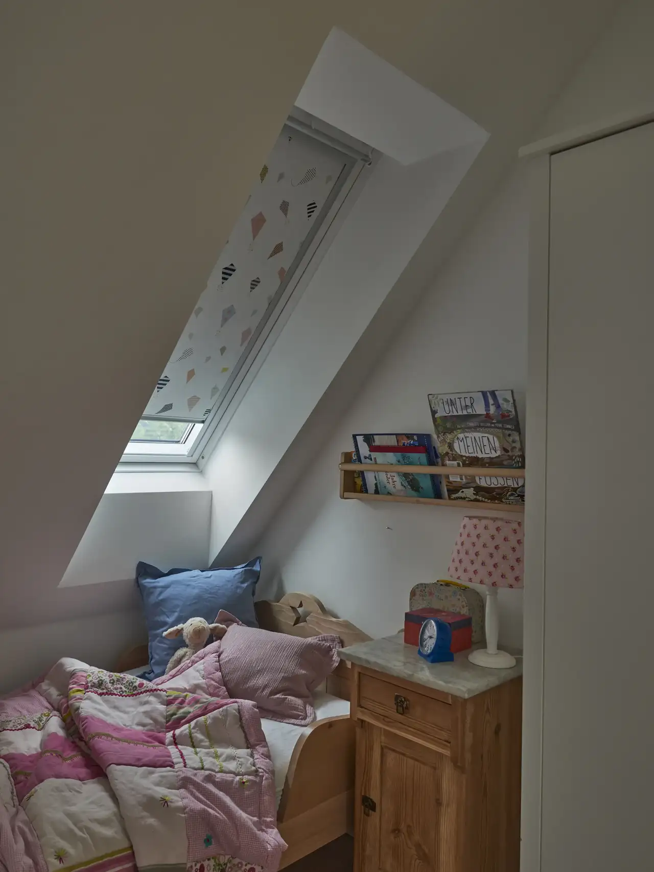 Una cameretta in mansarda: il letto è posizionato sotto una finestra per tetti con una tenda oscurante quasi del tutto chiusa.