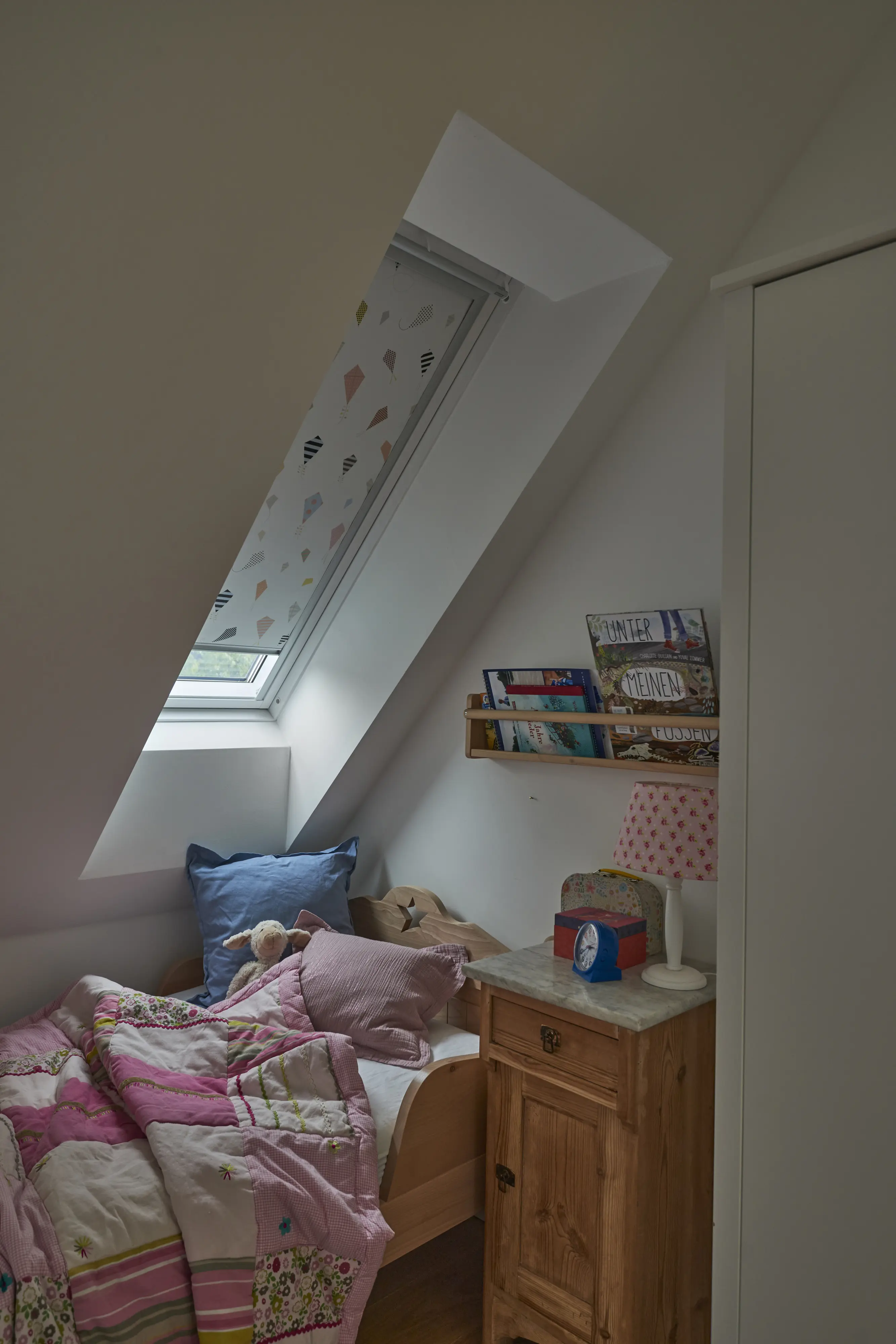 Chambre sous les combles : un lit placé sous une fenêtre de toit équipée d’un store occultant presque entièrement fermé.