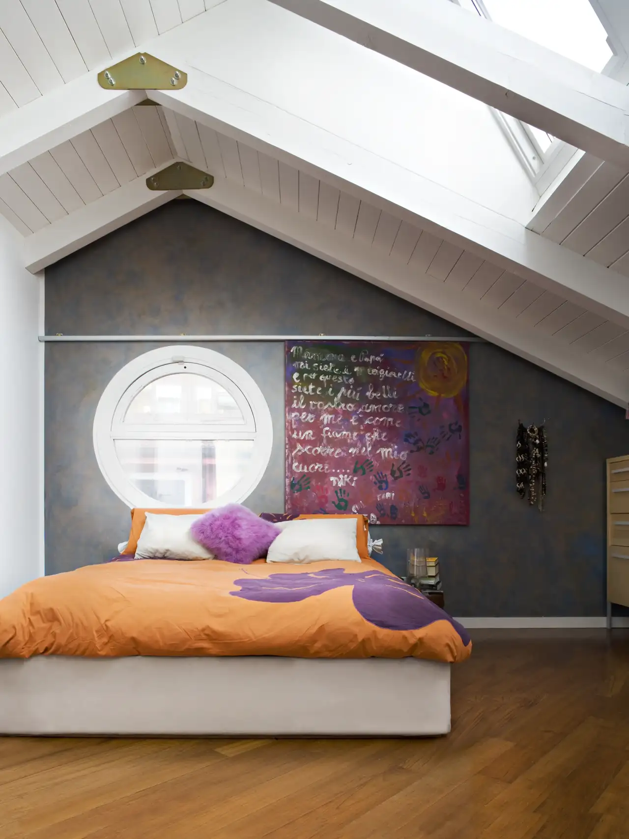 Chambre avec mur gris, lit orange et détails violets, éclairée par des fenêtres de toit.   Caption: Équilibrer couleurs vives et motifs rétro avec des lignes épurées et beaucoup de lumière naturelle : la clé d’un style années 80 revisité, frais et 