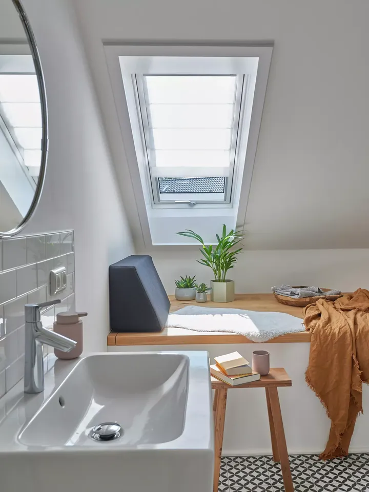 Modernes Badezimmer mit VELUX Dachflächenfenster, weißem Waschbecken, Pflanzen und hölzernem Hocker mit Büchern.