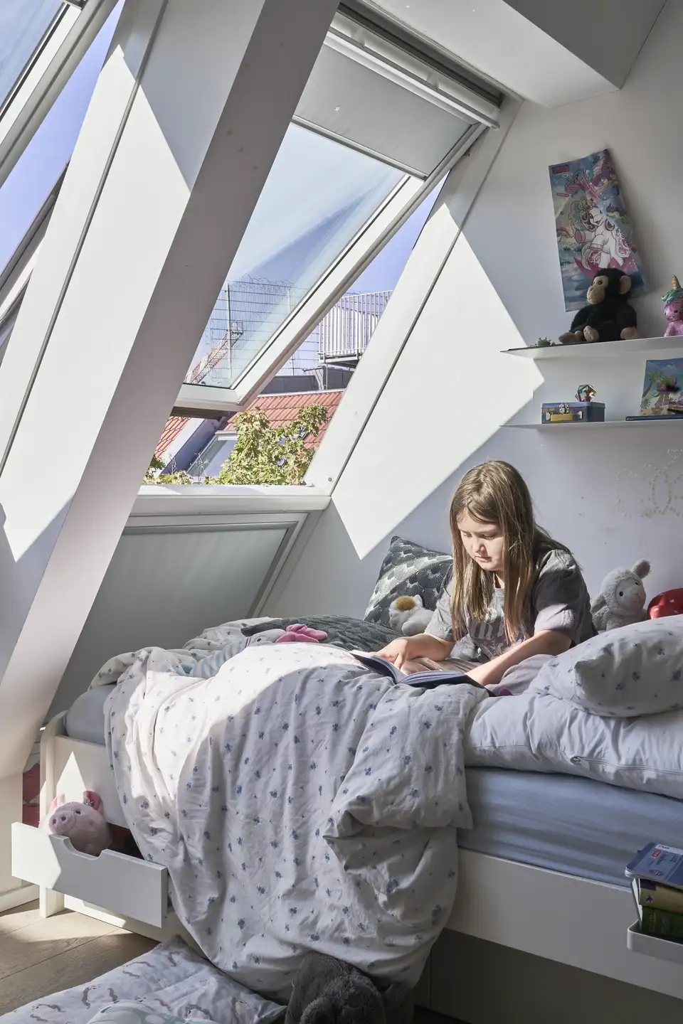 Dievčatko číta na posteli pod veľkými strešnými oknami a užíva si prirodzené svetlo v podkrovnej spálni. | VELUX