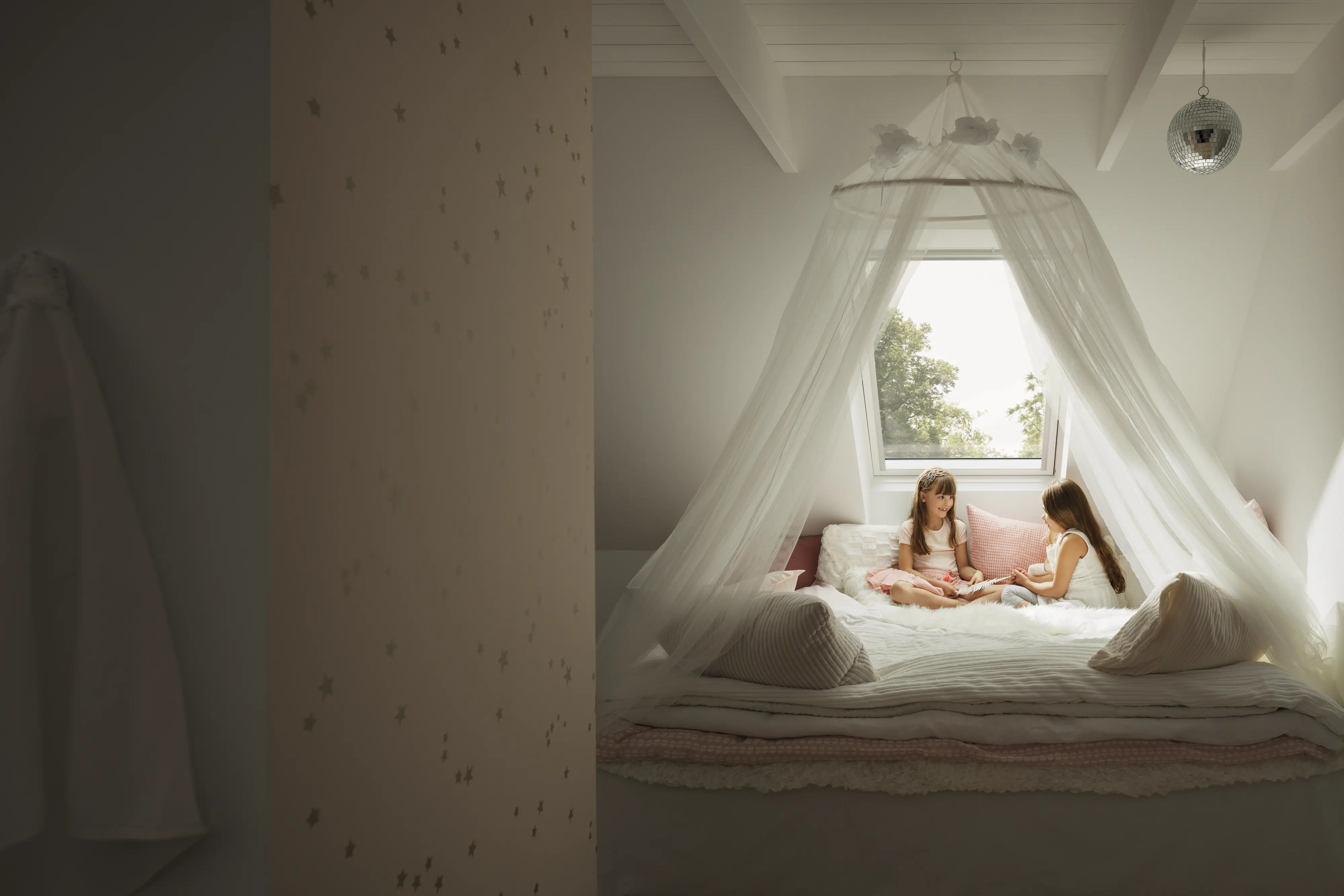 Chambre d’enfants sous combles avec fenêtre de toit VELUX, deux fillettes jouant sur un lit à baldaquin. 