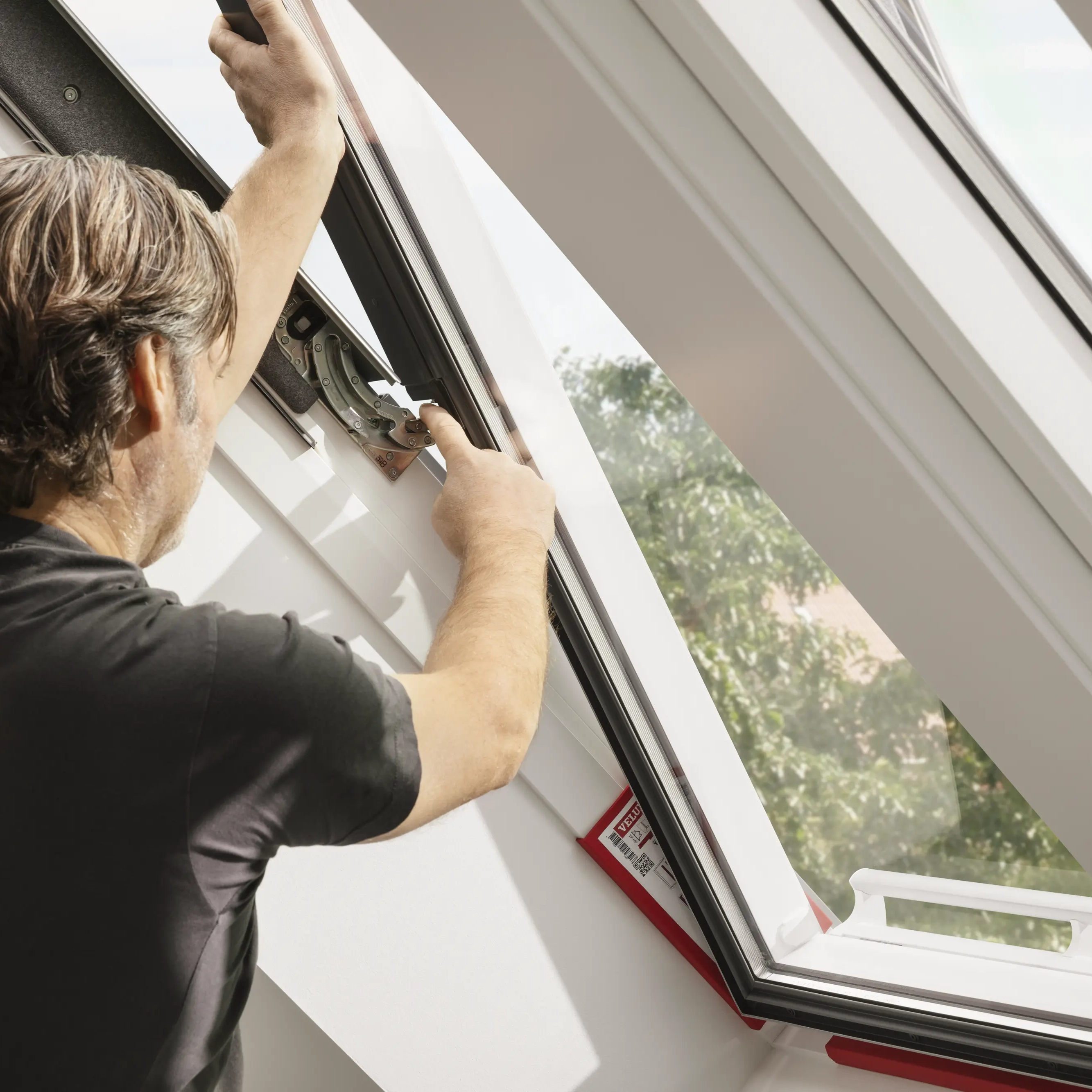 Man adjusting an open VELUX roof window in a bright attic room.