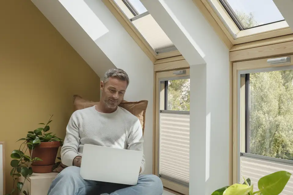 Férfi, aki a nagy VELUX tetőablakok mellett ül egy széken, és a laptopozik.