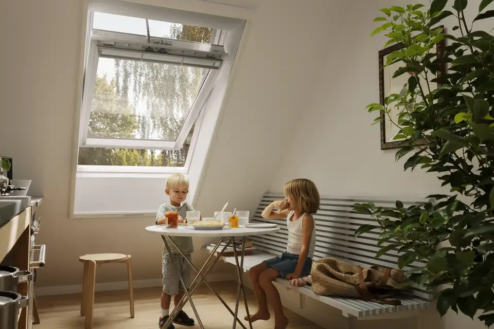 Twee kinderen eten hun tussendoortje in een keuken bij een VELUX dakvenster met een muggengaas.