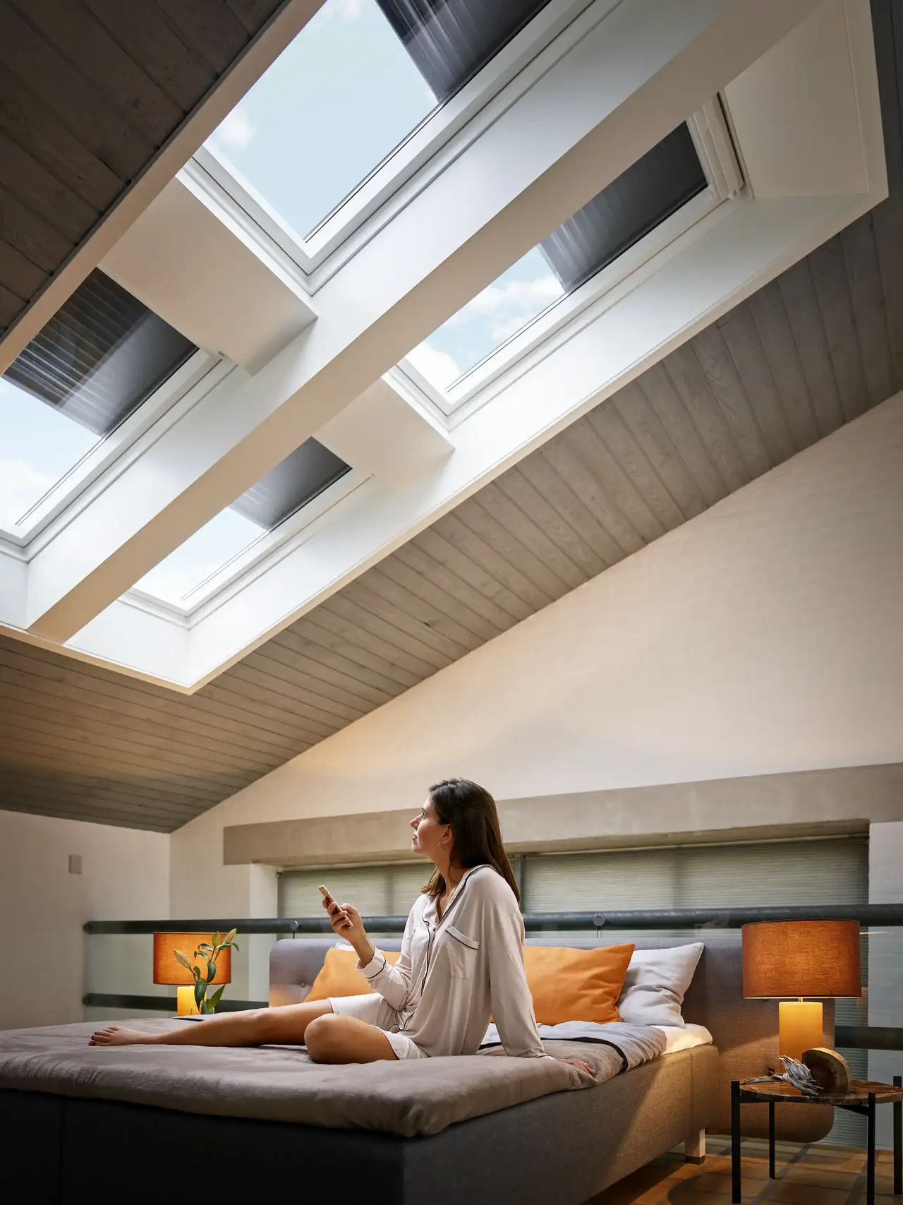 Yatakta oturan bir kadın, VELUX App Control uygulamasıyla VELUX çatı pencerelerini ve dış panjurları kontrol ediyor 
