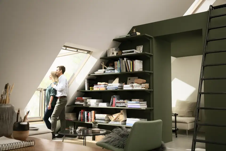 Mansarda open space con grande libreria aperta in alto a separare due ambienti, entrambi illuminati da finestre per tetti