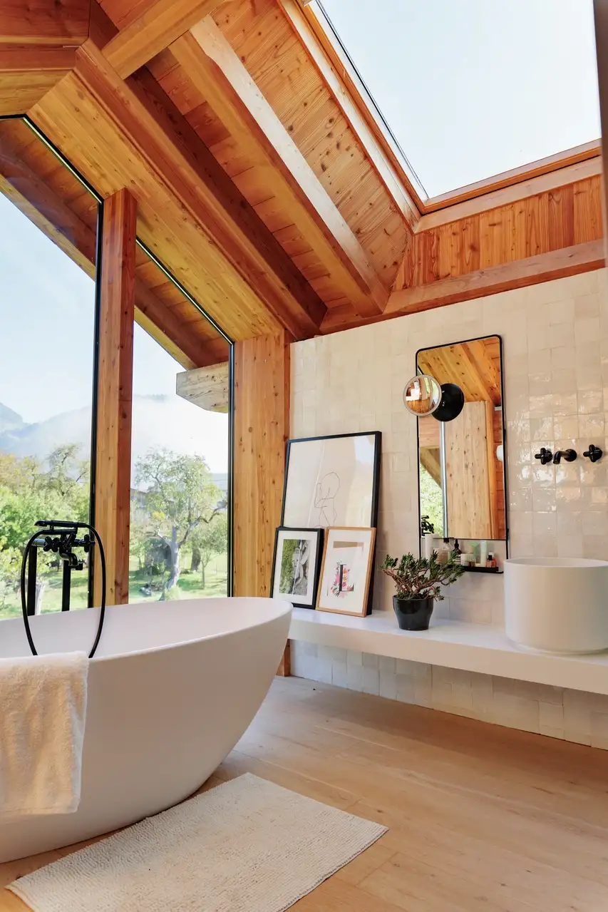 Une salle de bain lumineuse avec une baignoire blanche autoportante, de grandes fenêtres encadrées de bois avec vue sur la verdure, une fenêtre de toit et un comptoir minimaliste avec des décorations et des œuvres encadrées.
