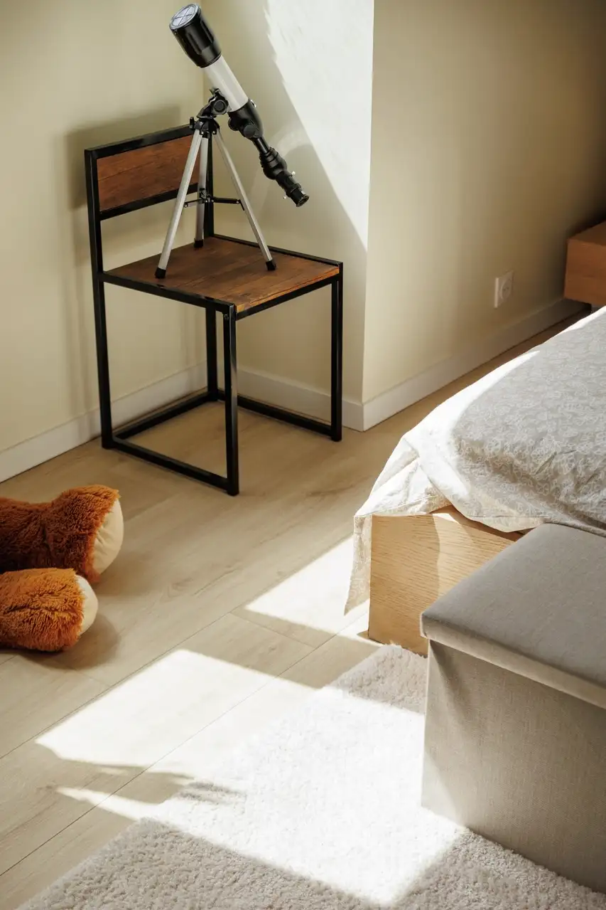 : Un télescope posé sur une chaise dans un coin de chambre, baigné de douce lumière, à côté d'un lit confortable. 