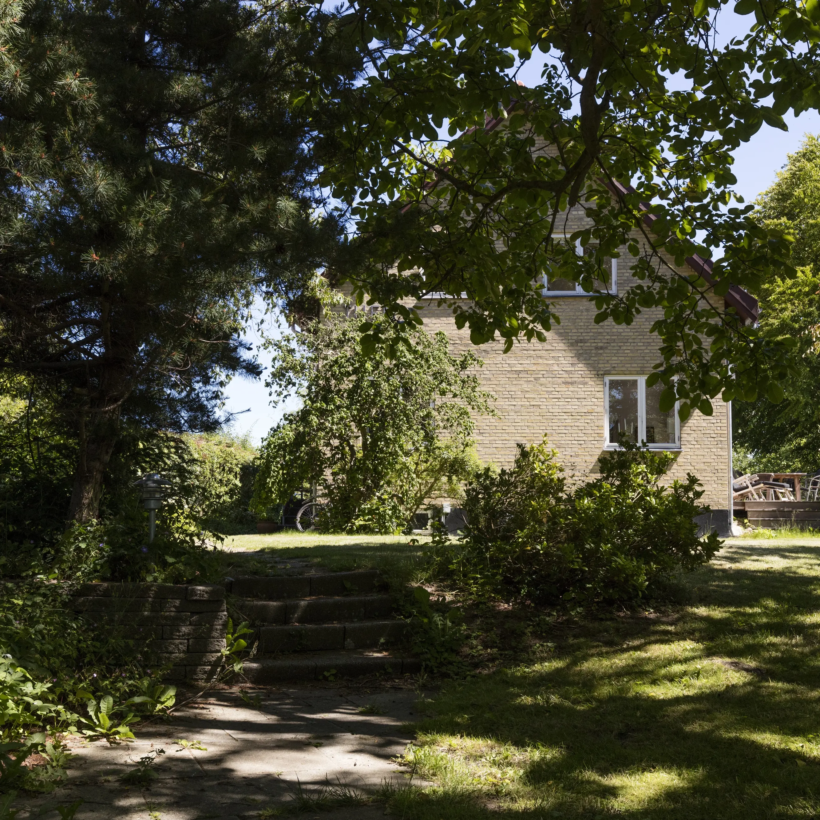 Une maison individuelle en briques se trouve au centre d’un jardin arboré, avec de grands arbres offrant de l’ombre naturelle et un espace extérieur verdoyant baigné de lumière estivale.