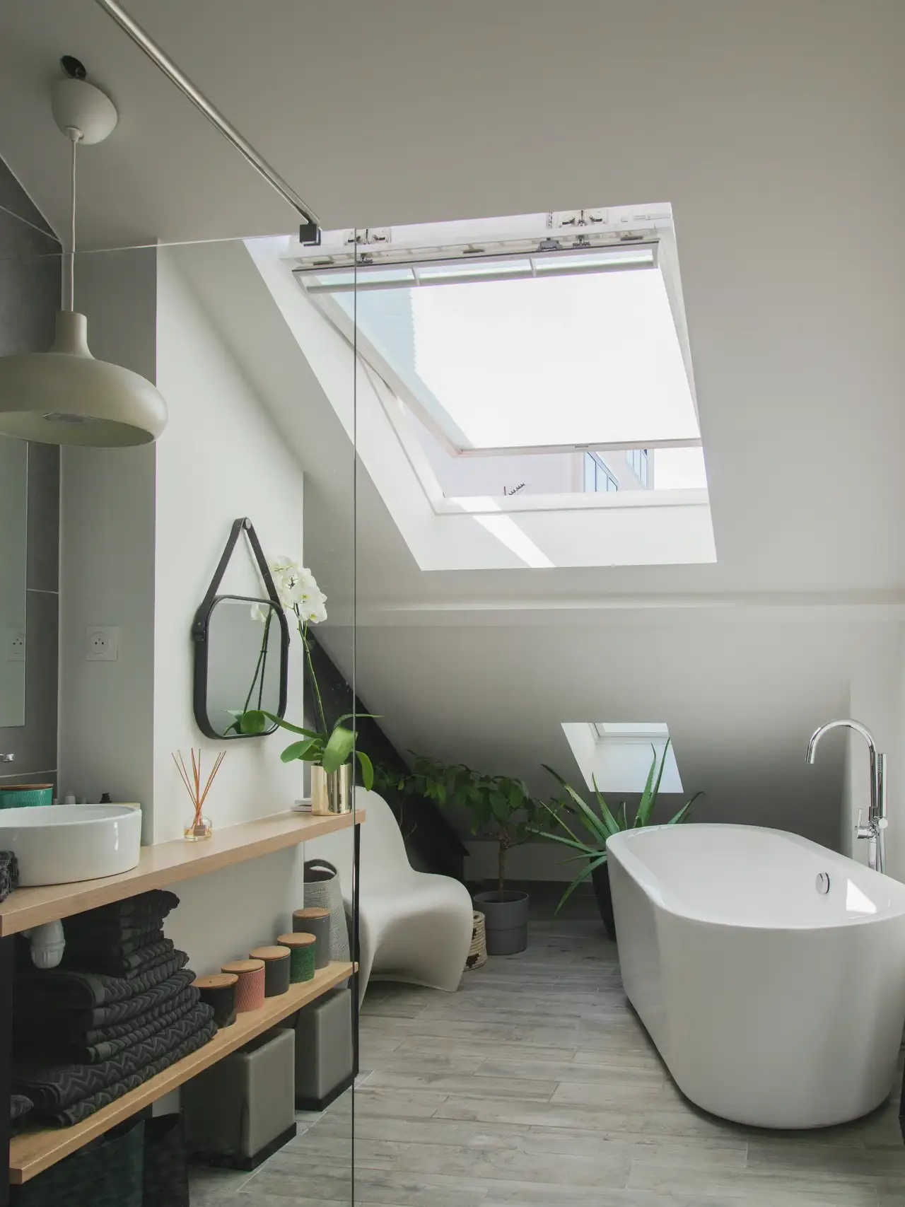 Une salle de bain moderne avec une lumière naturelle entrant par une fenêtre de toit ouverte. La pièce comprend une baignoire autoportante, un lavabo avec un miroir rond au-dessus et diverses plantes apportant de la verdure. Le design est minimaliste avec des couleurs neutres, et la fenêtre de toit ajoute un élément architectural intéressant qui améliore la luminosité de l’espace.