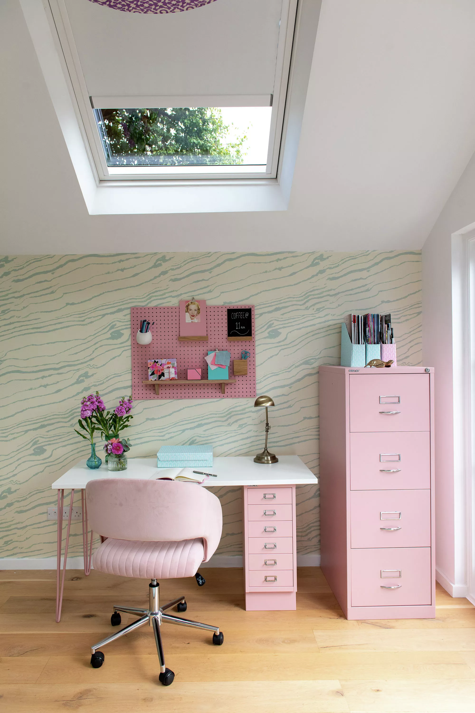 Chambre lumineuse avec Velux, bureau rose et chaise assortie, décorée d’éléments artistiques et d’une plante.