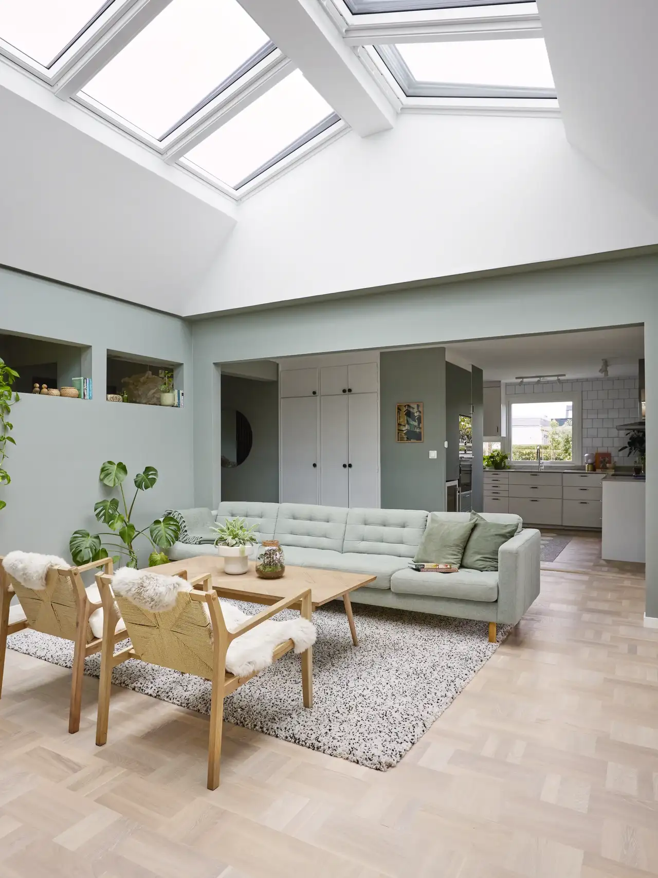 Lys og moderne innredet stue med grønne vegger, lysegrå sofa og tremøbler. Dagslyset som flommer inn gjennom de store takvinduene, skaper en åpen atmosfære.