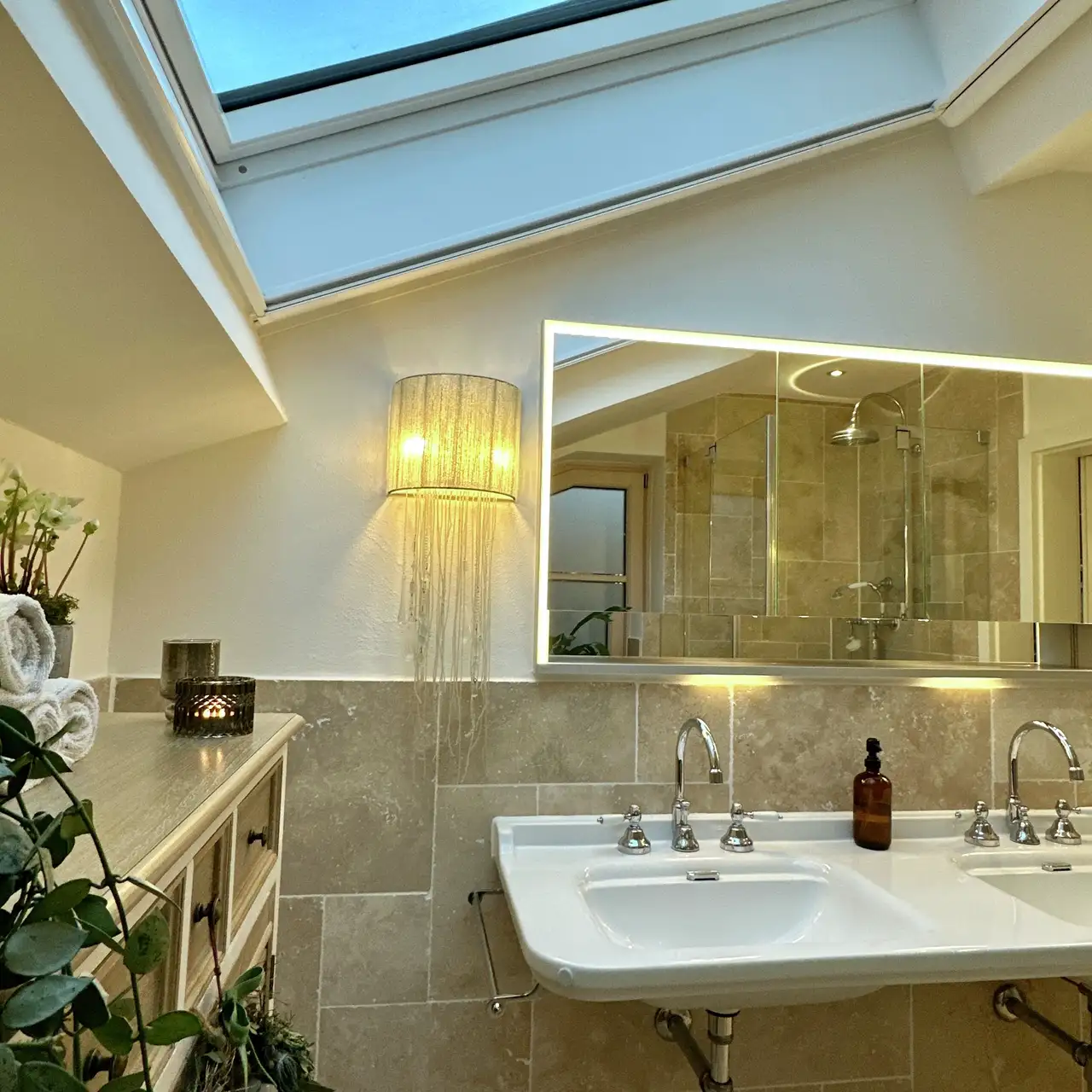 Salle de bain sous les combles avec double vasque, miroir rétroéclairé, applique décorative et fenêtre de toit. 