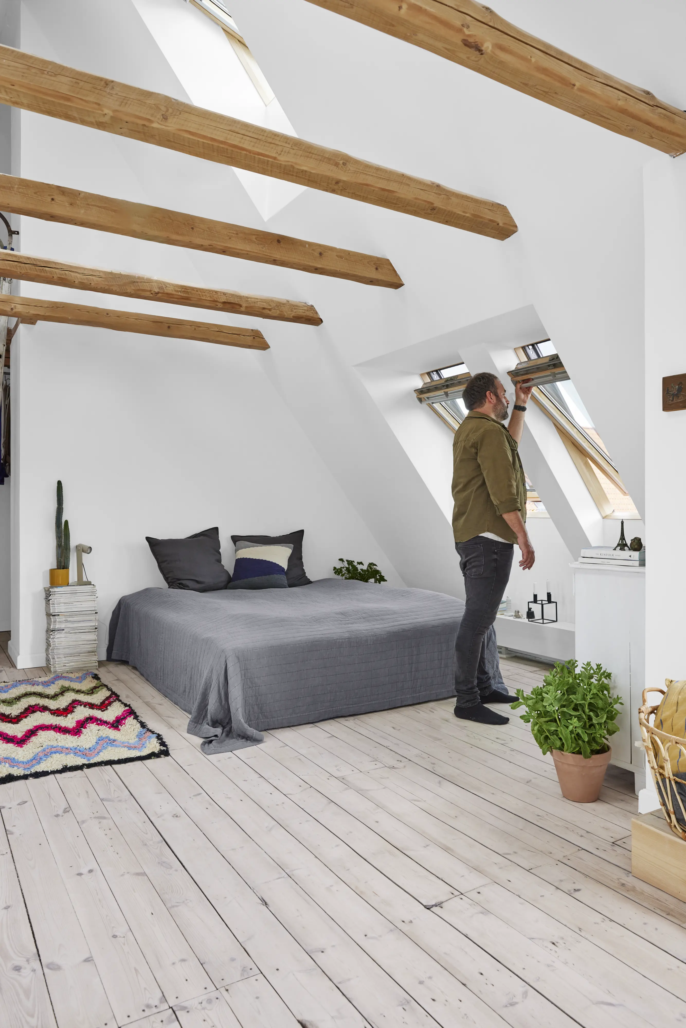 Vīrietis atver jumta logu gaišā guļamistabā ar atsegtām koka sijām. | VELUX