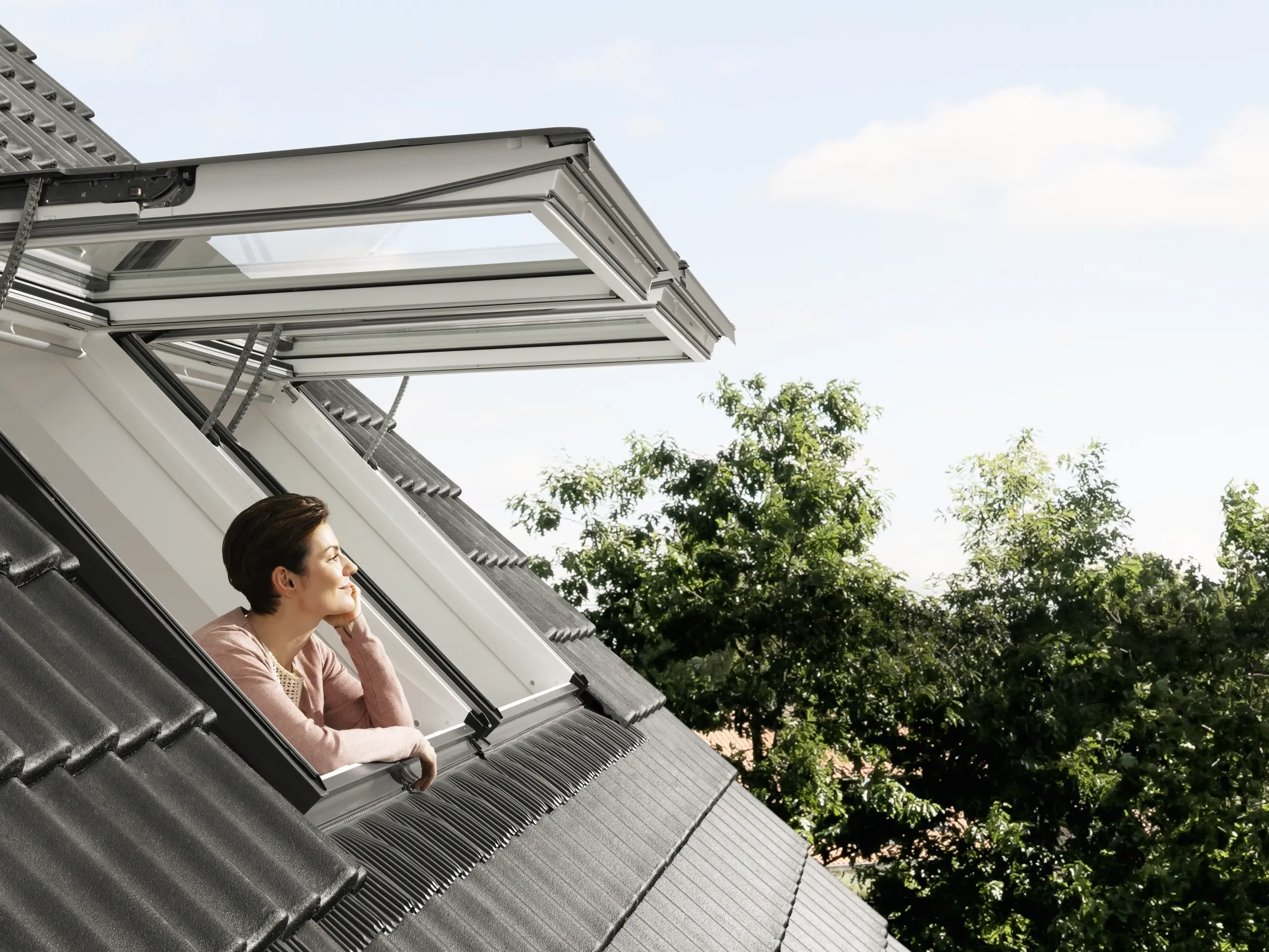 Atvērts VELUX jumta logs ar skatu uz kokiem un debesīm.