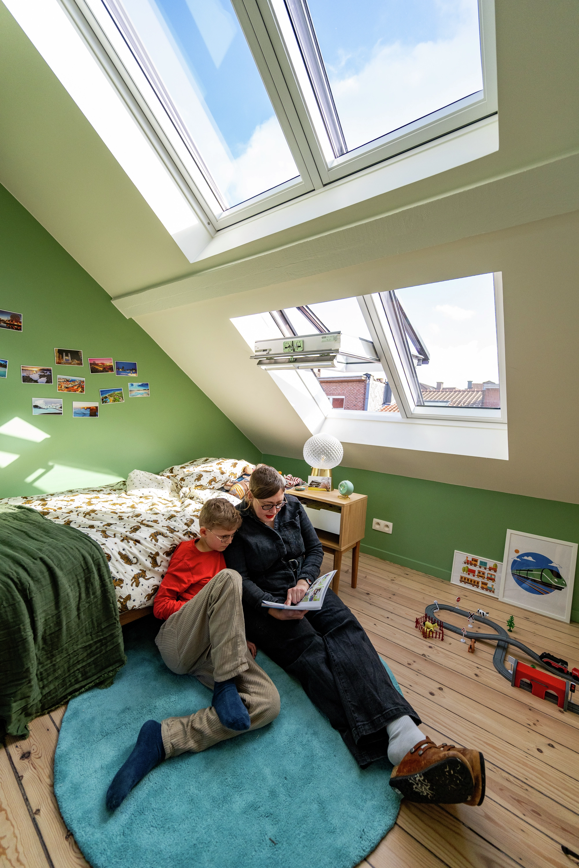 Un garçon lit avec sa maman dans sa chambre sous les toits, éclairée par une combinaison de fenêtres de toit. 